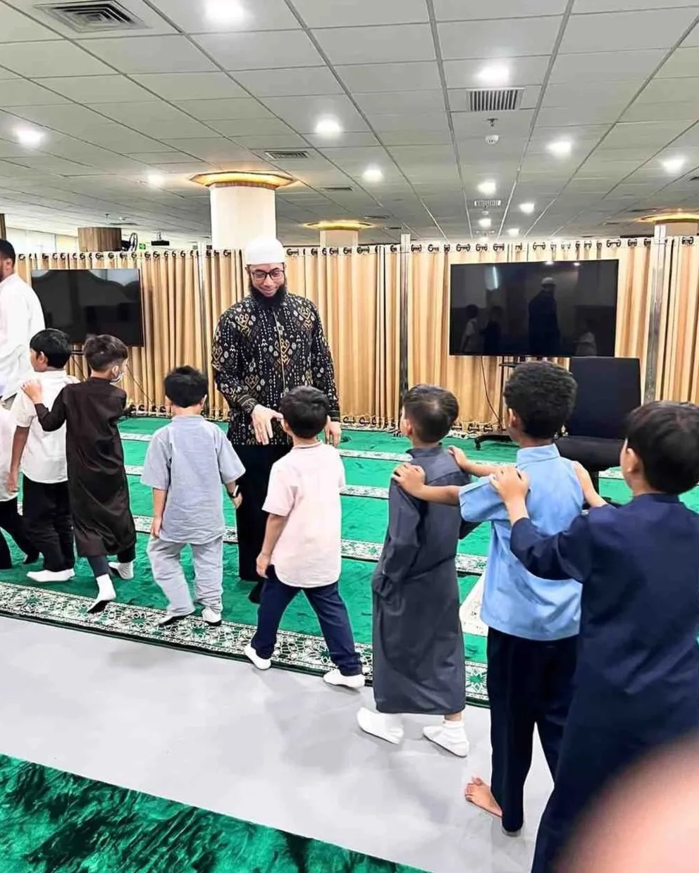 A man in traditional Islamic dress leading young boys in a prayer activity inside a mosque or prayer room. The boys are standing in a line, following the man, with some touching each other's shoulders. The room has a green carpet, curtains, and large monitors on the walls.