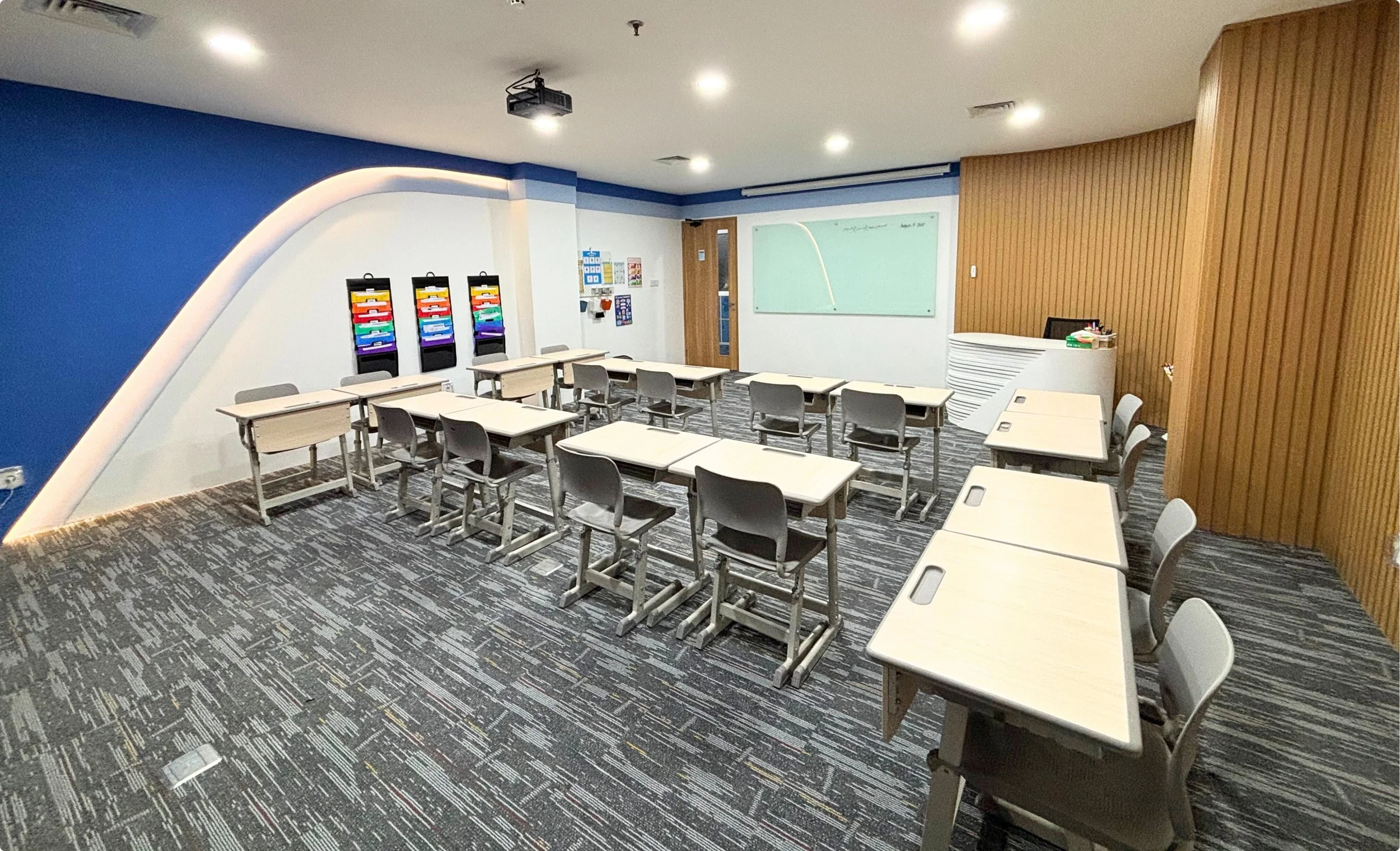 Empty classroom with desks and chairs, whiteboard, colorful materials, wooden wall, ceiling lights, and a gray patterned carpet.