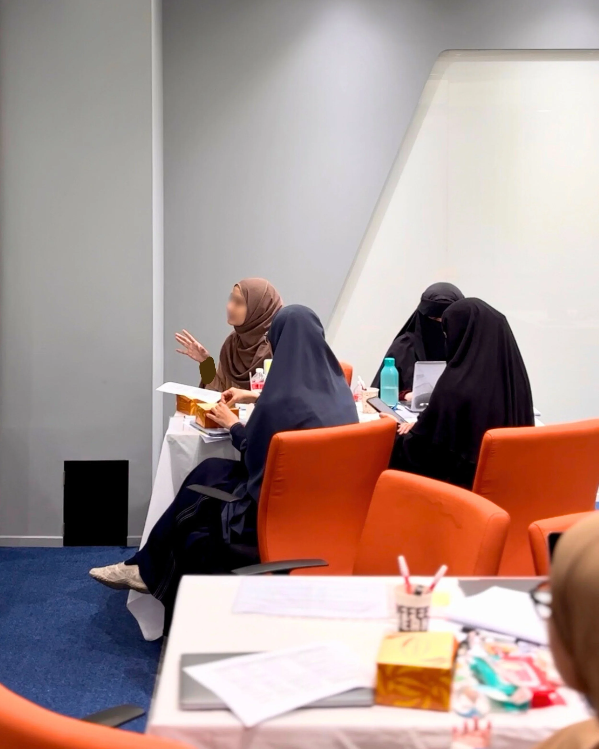 Four women wearing hijabs sitting at a conference table in a modern office, engaged in discussion, with various documents, water bottles, and handouts on the table.