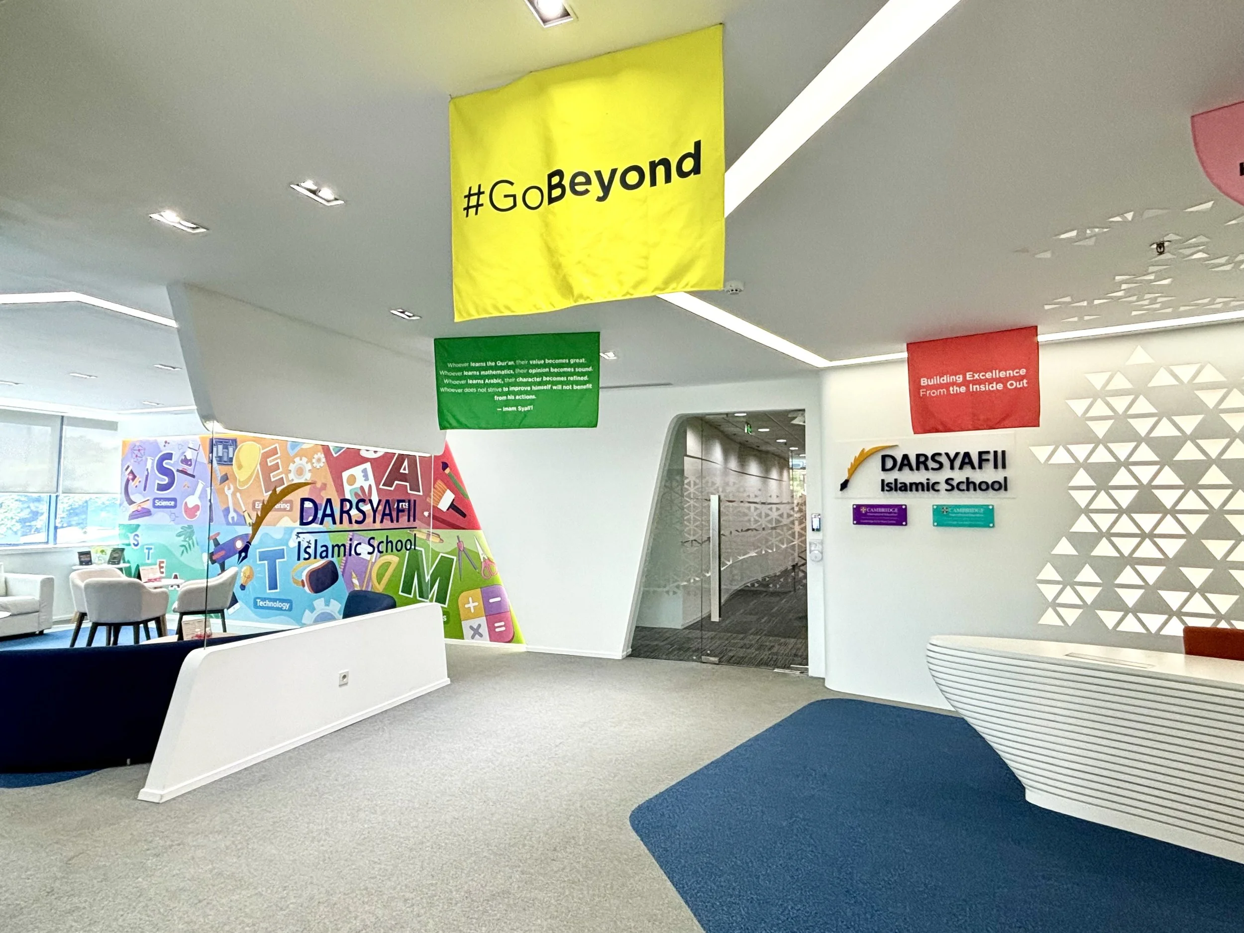 Interior of Daryafi Islamic School with colorful wall art and signage, yellow and red banners hanging from ceiling, white walls, modern furniture, and an open doorway.