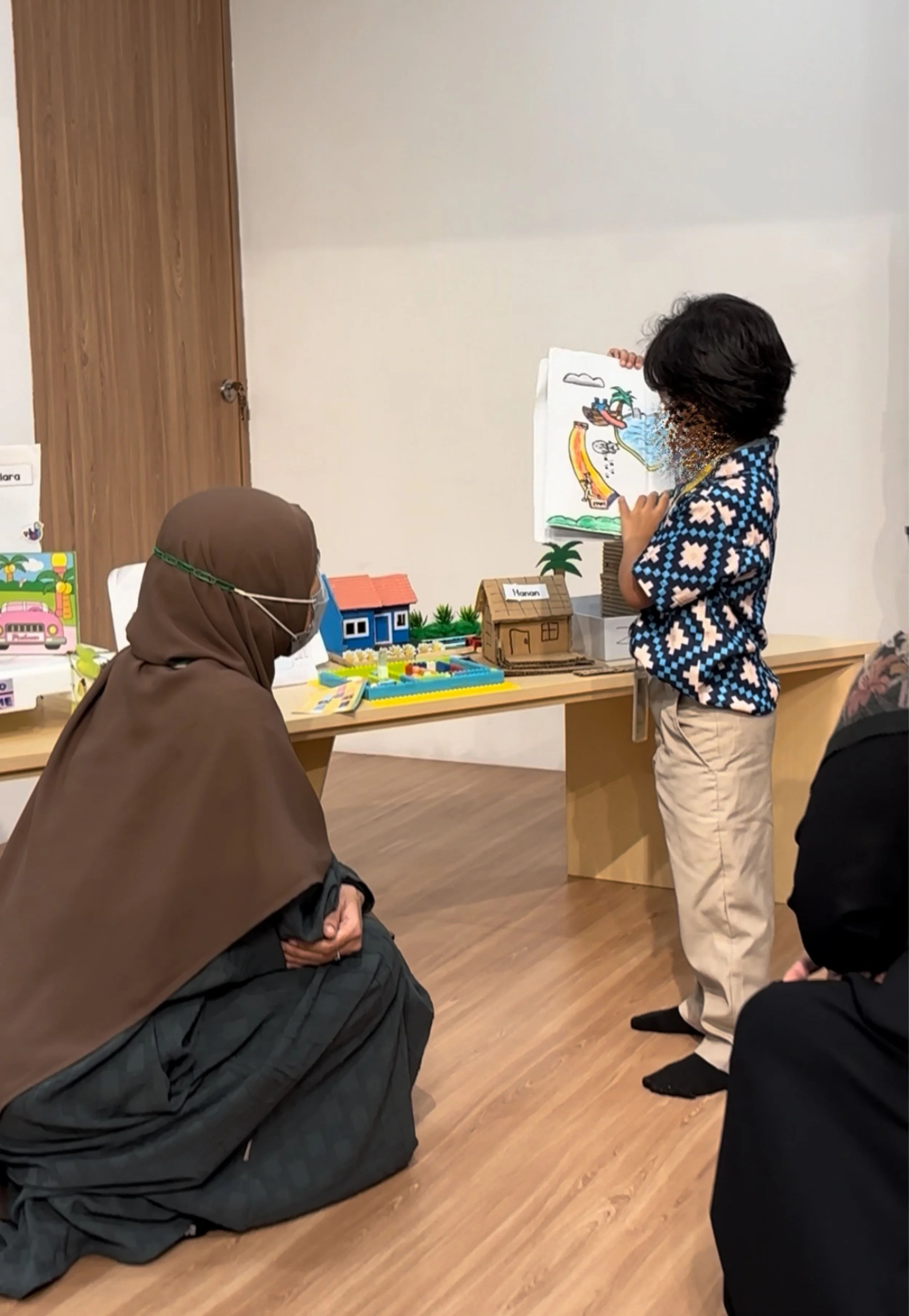 A young boy presenting a drawing of a tropical island scene to an adult woman kneeling on the floor, with other display items and miniature models on a table in the background.