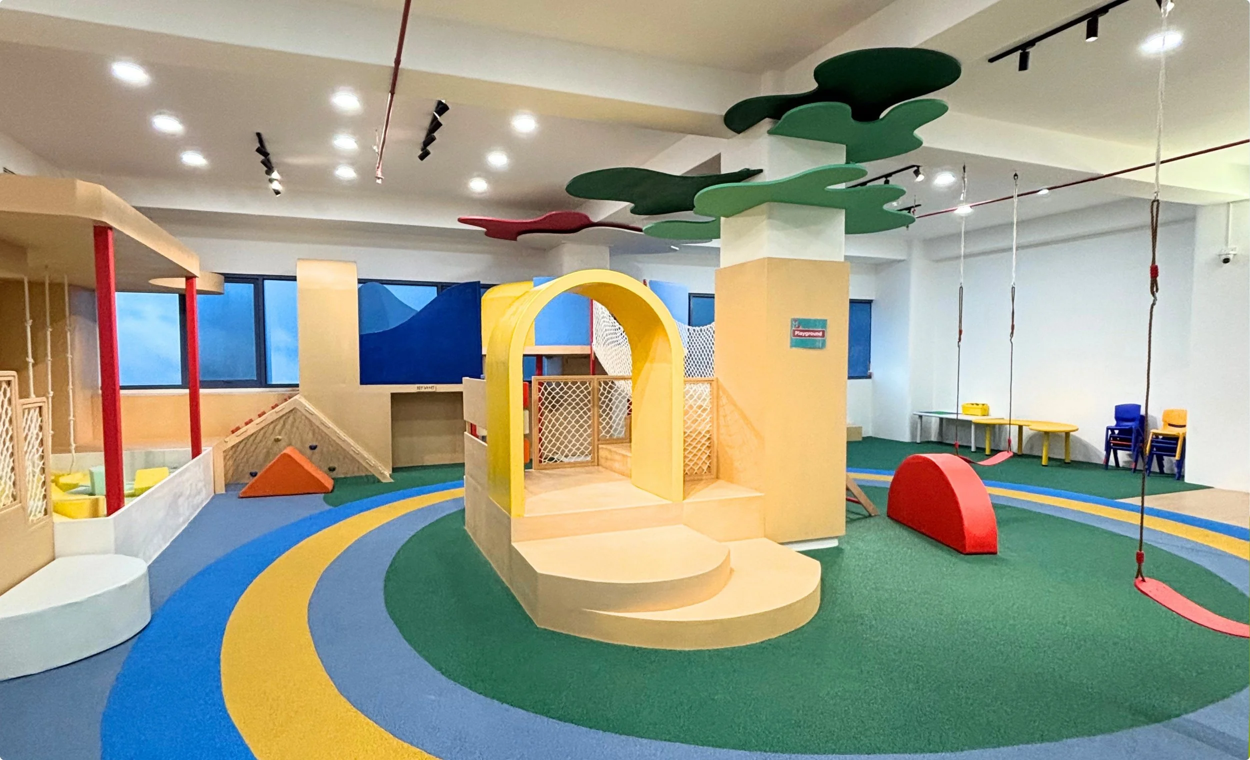 Indoor children's playground with colorful play structures, swings, and a rainbow-patterned carpet.