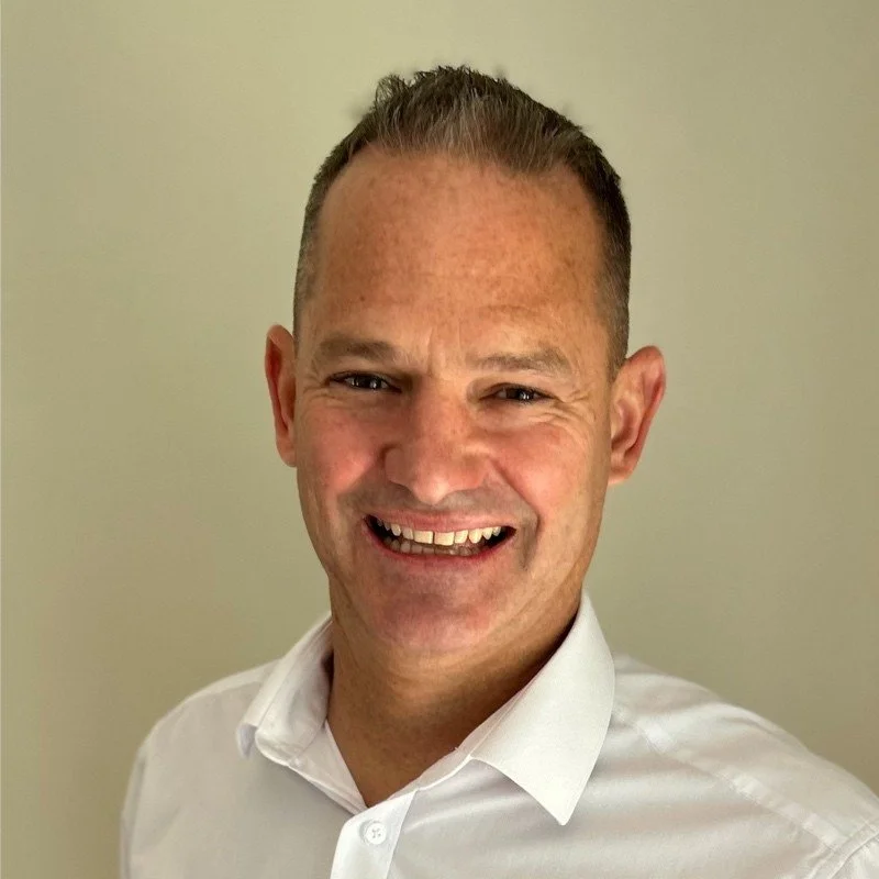 A smiling man with short hair wearing a white collared shirt against a plain background.