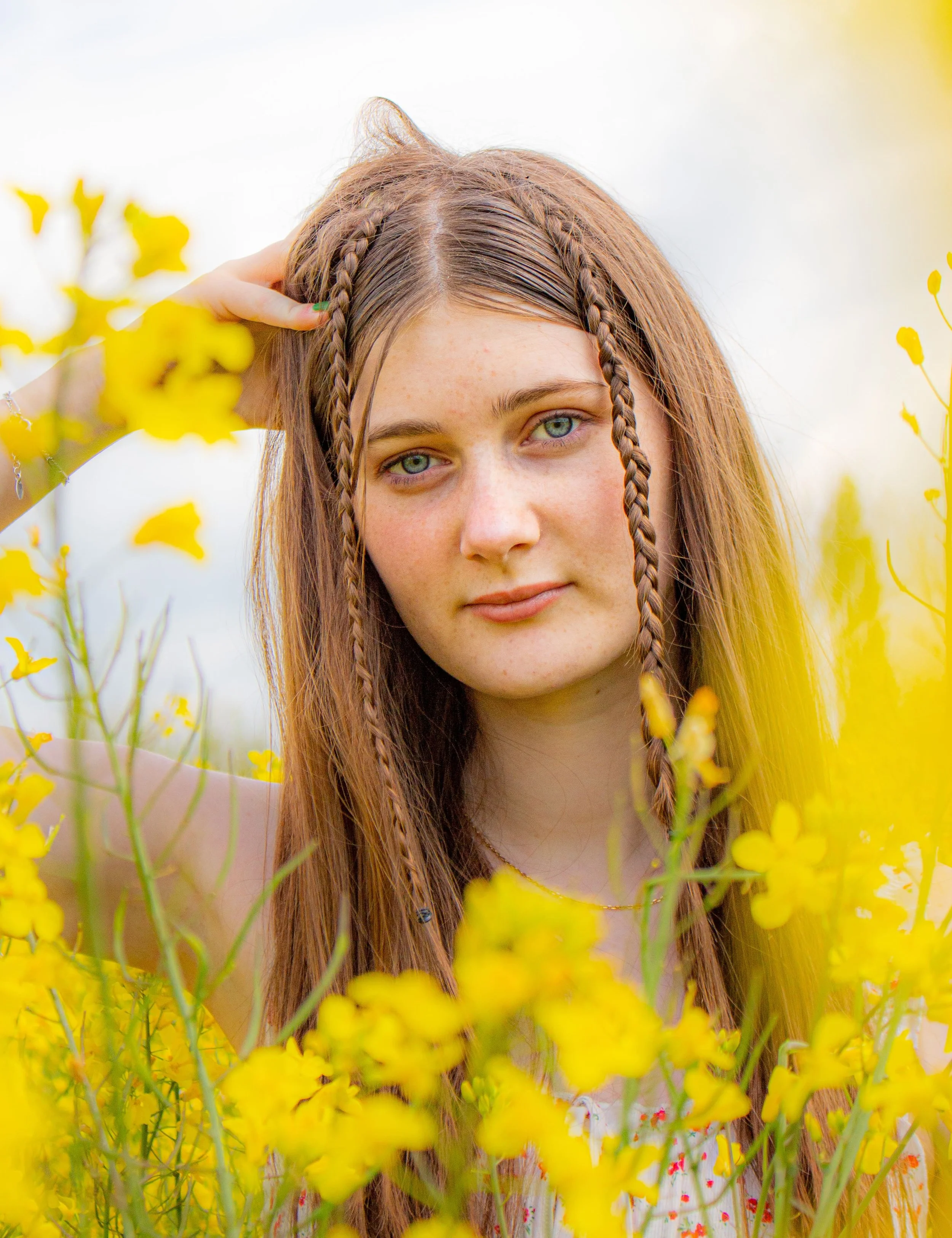 Jeune femme aux cheveux longs et bruns avec deux tresses, dans un champ de fleurs jaunes, regardant l'objectif.