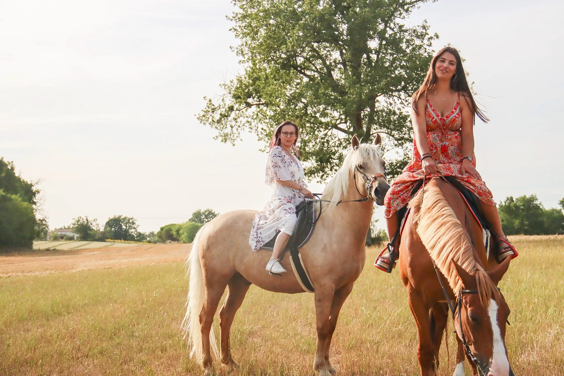Deux jeunes femmes chevauchent des chevaux dans un champ large avec un arbre en arrière-plan sous un ciel en partie nuageux.