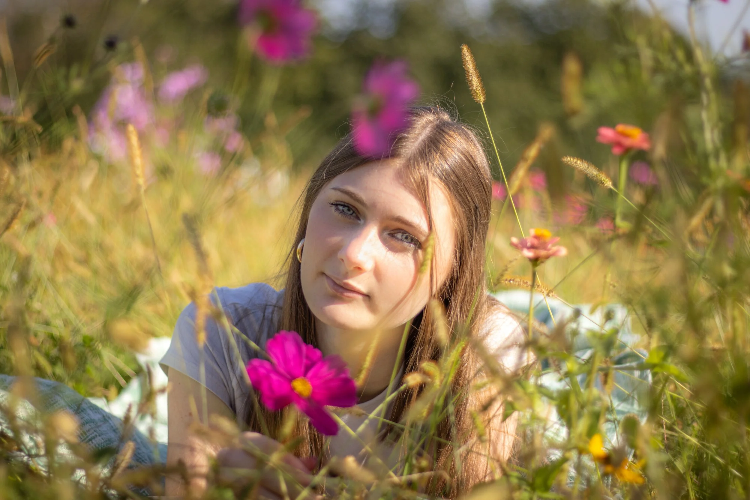 Une jeune femme aux cheveux bruns et yeux bleus, allongée dans un champ de fleurs coloration rose et jaune, sous un soleil brillant.