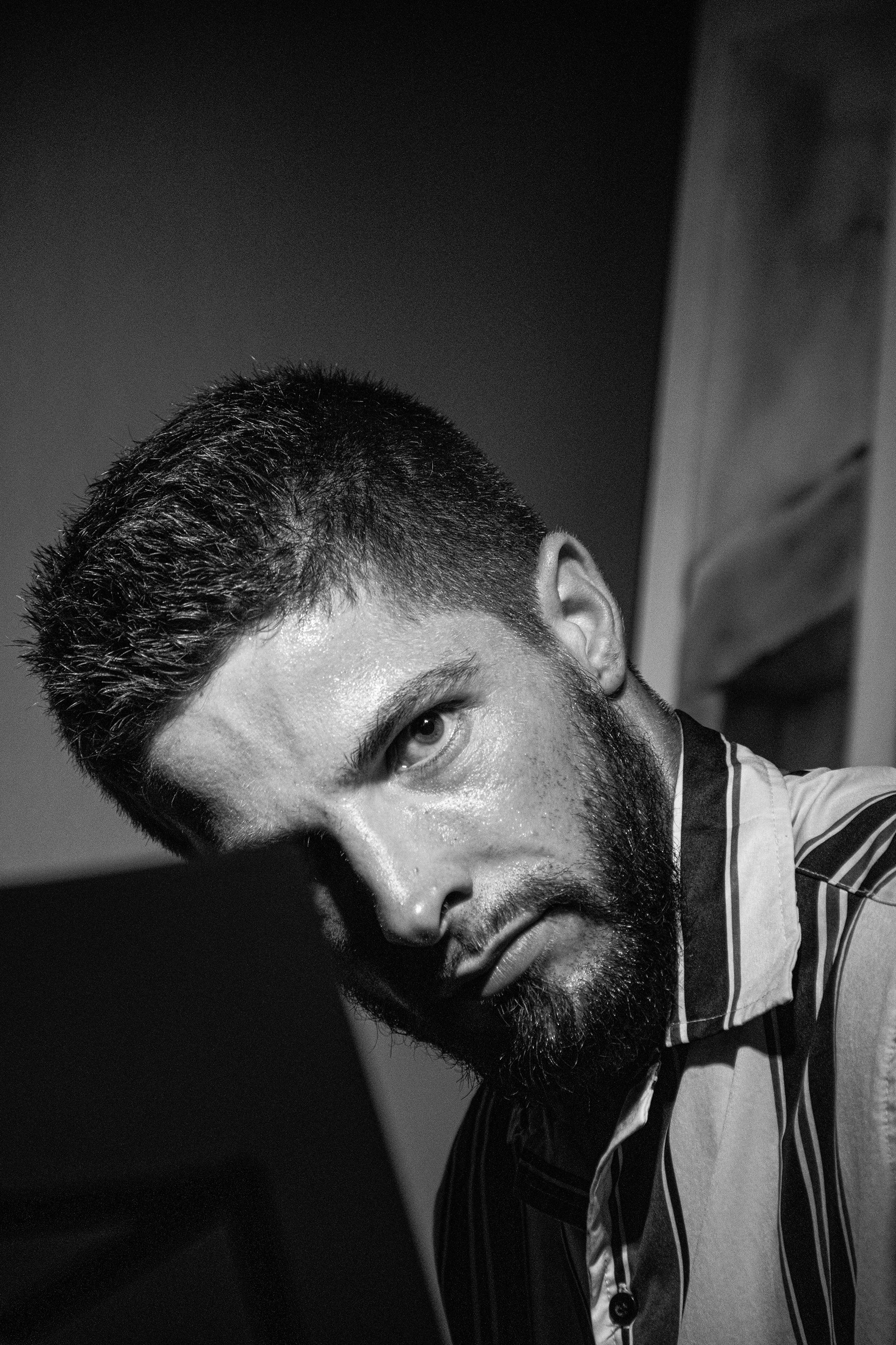Un homme avec barbe et cheveux courts, portant une chemise à rayures, regarde intensément l'objectif en étant penché en avant, dans un environnement intérieur. L'image est en noir et blanc.