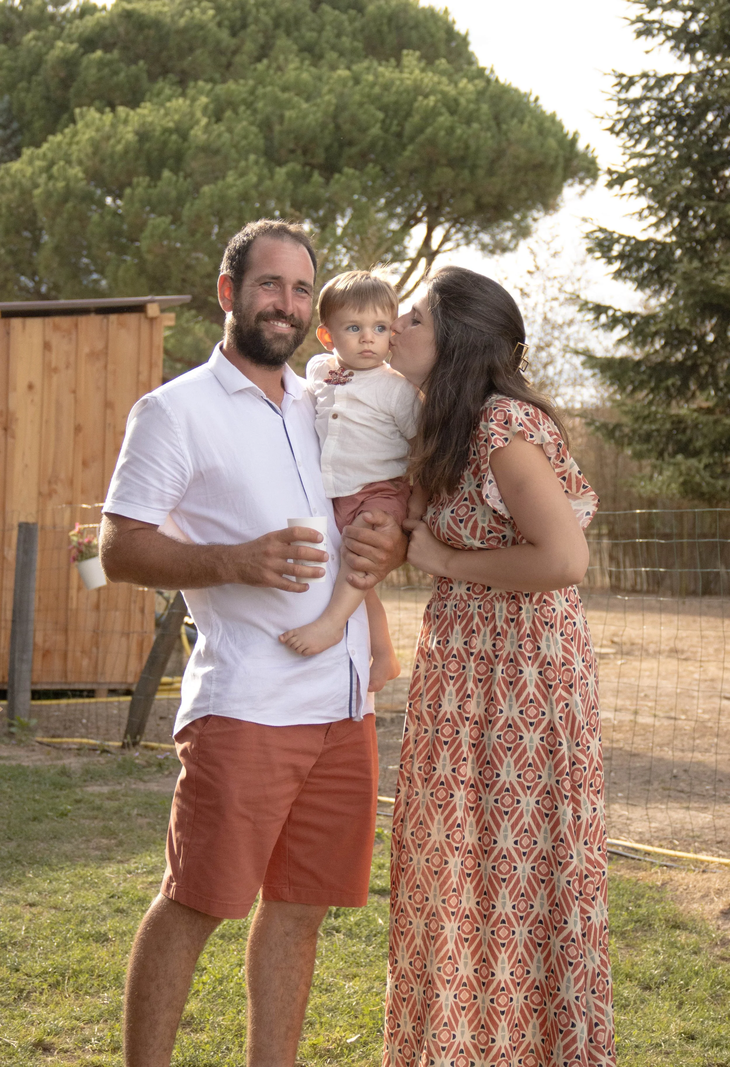 Une famille pose ensemble dans un jardin, avec un homme souriant, une femme embrassant un enfant, tous sous un ciel clair