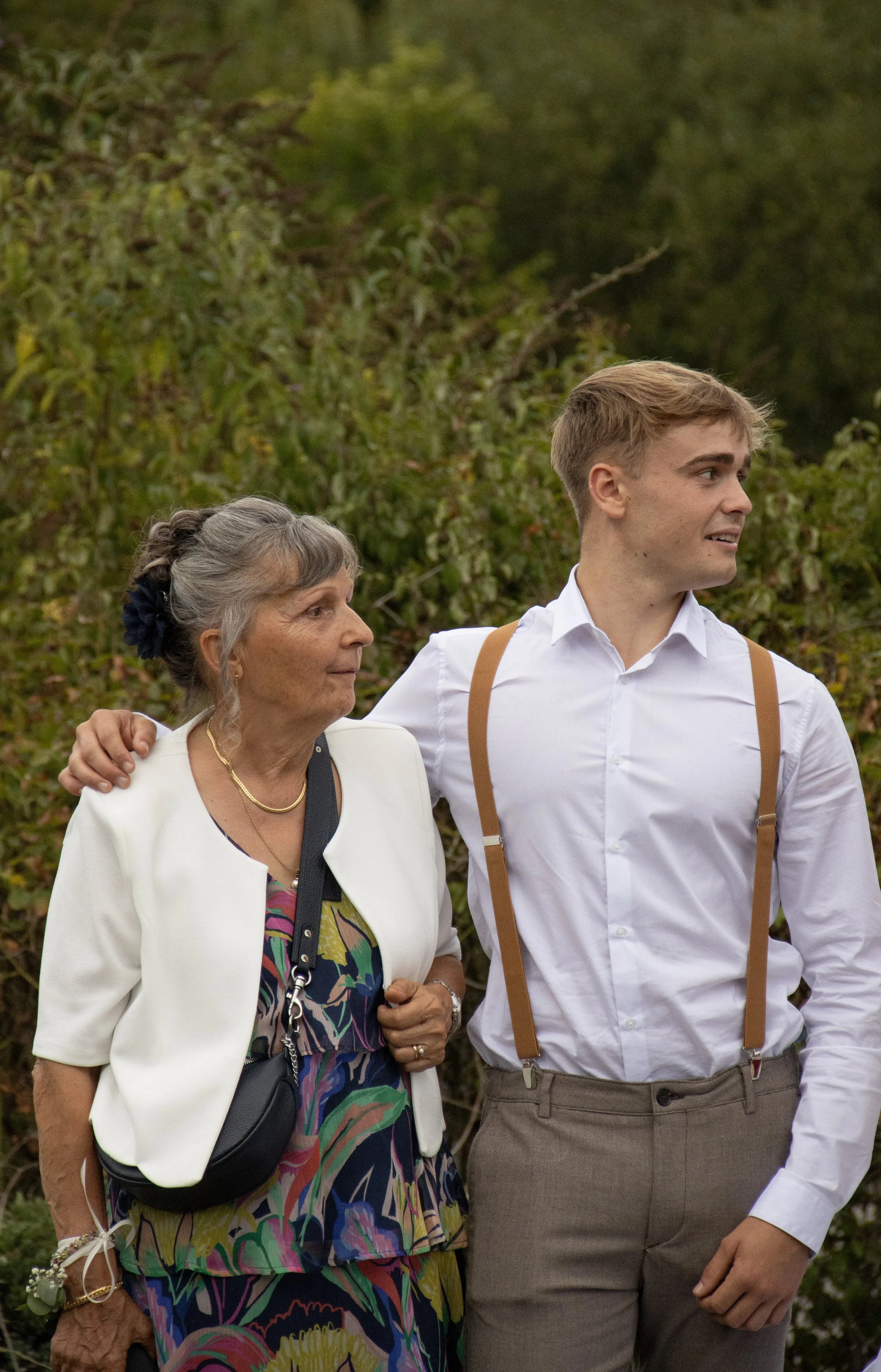 Une femme âgée et un jeune homme se tiennent l'un à côté de l'autre dans un extérieur verdoyant, la femme avec une main sur l'épaule du jeune homme, tous deux regardant dans la même direction.