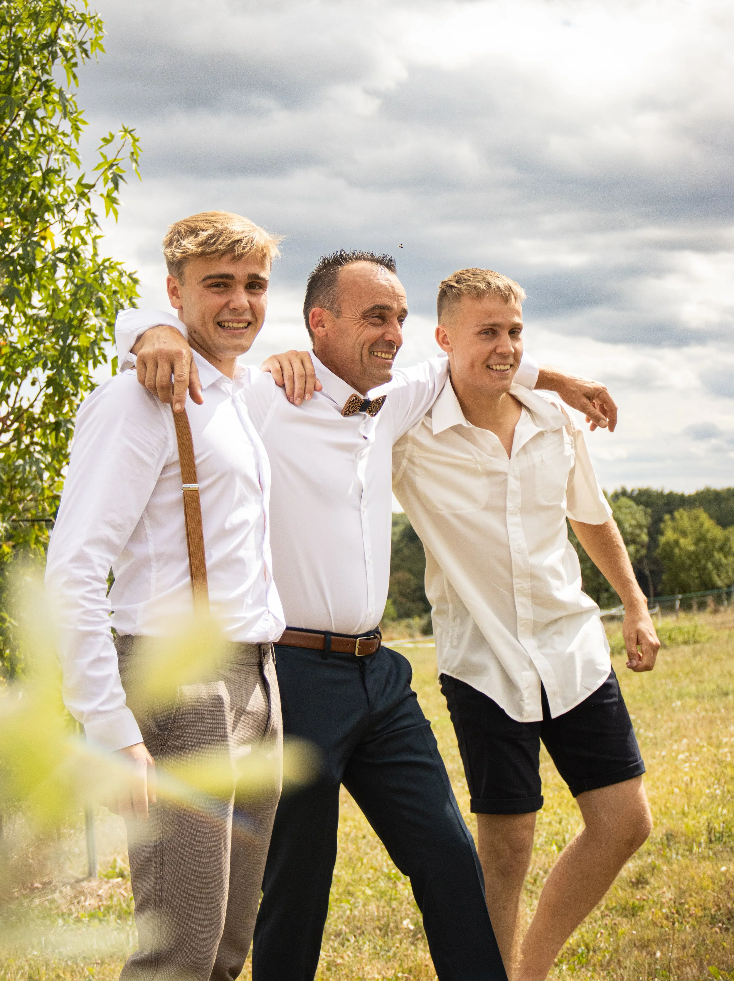 Trois hommes souriants marchant ensemble dans un champ lors d'une journée ensoleillée, entourés de verdure.