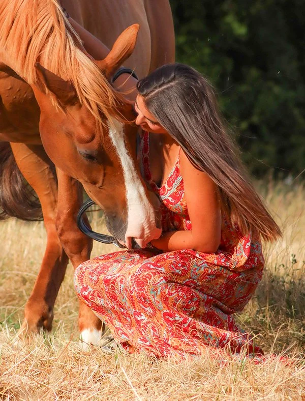 Une femme en robe colorée s'agenouille dans un champ, caressant un cheval brun avec une ligne blanche sur le visage.