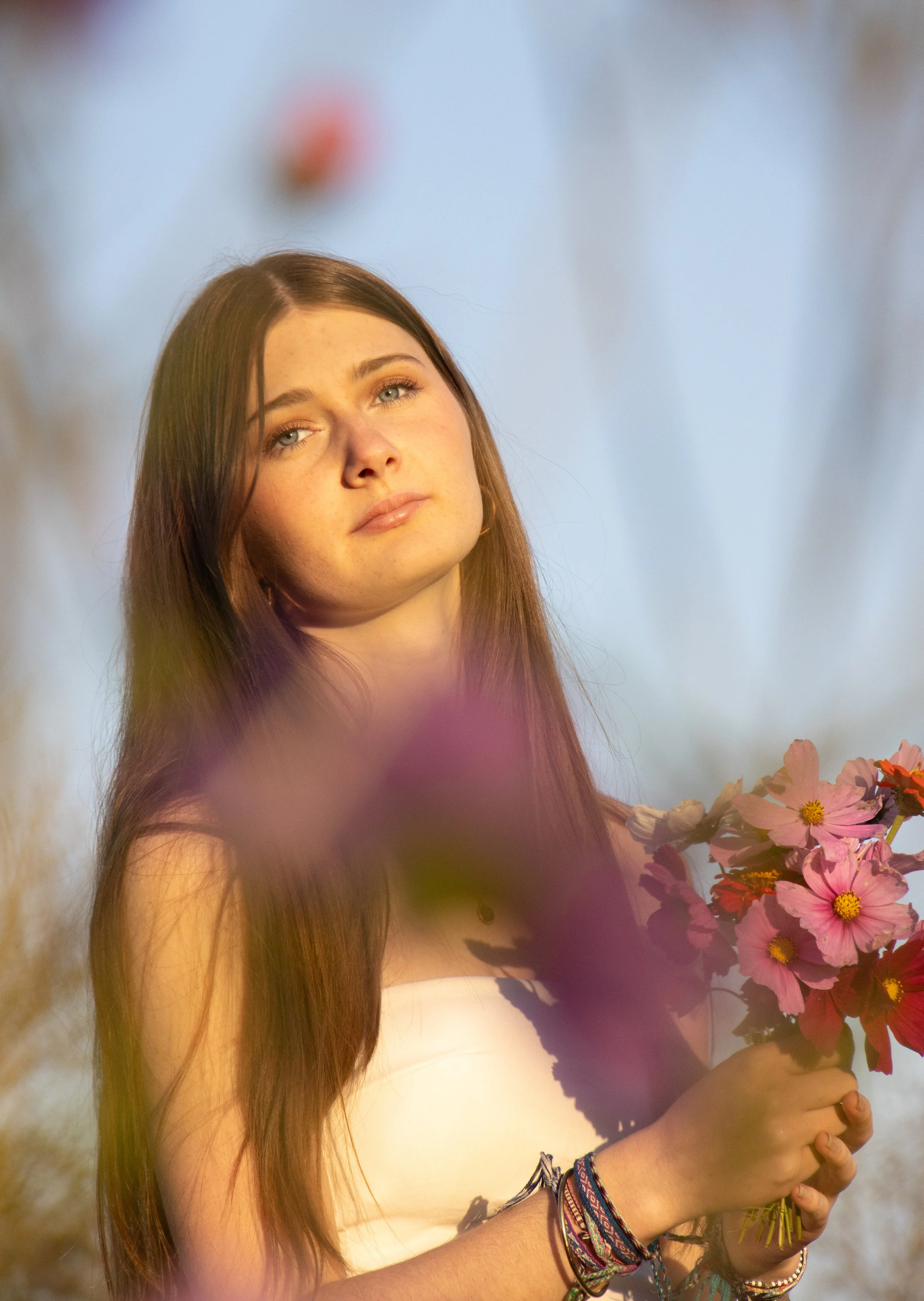 Une jeune femme aux cheveux longs, tenant un bouquet de fleurs roses et rouges, regarde vers le haut avec une expression douce, lors d'une séance photo avec un fond flou de nature.