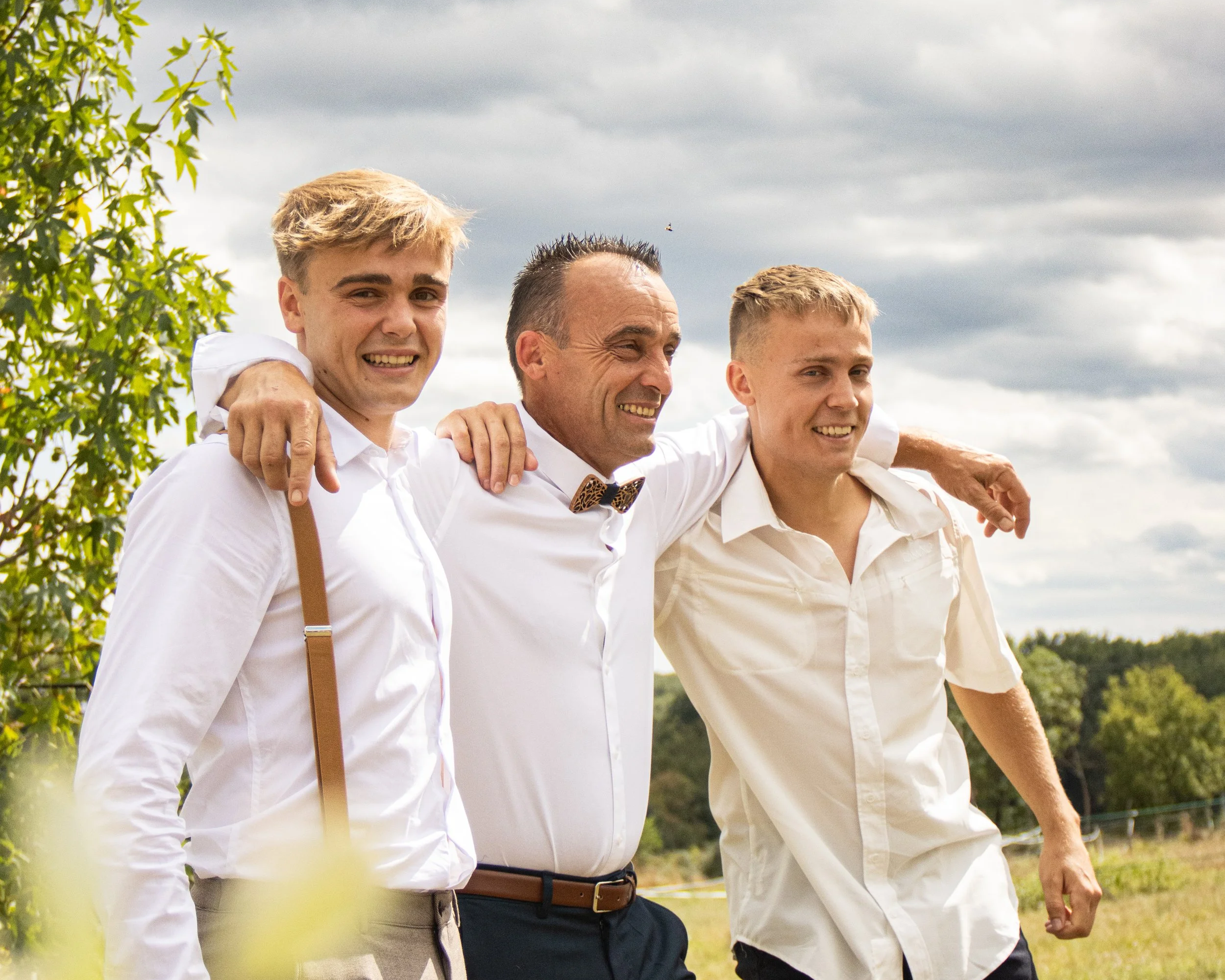 Trois hommes souriants en chemises blanches, avec leurs bras autour des épaules, marchant dans un champ avec un ciel nuageux en arrière-plan.