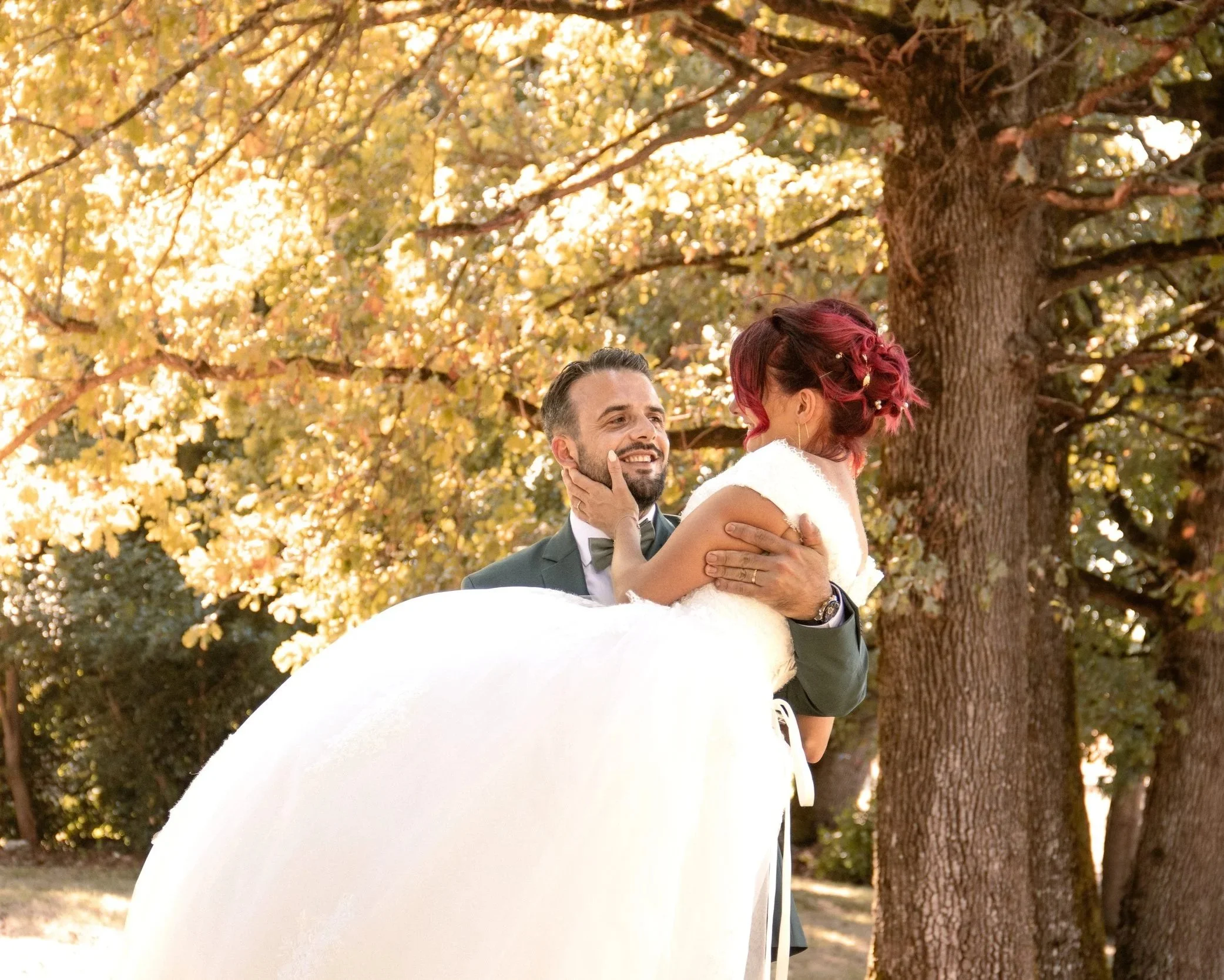 Un homme en costume noir tient une femme en robe de mariée blanche dans ses bras devant un arbre avec des feuilles dorées en automne, ils semblent heureux.