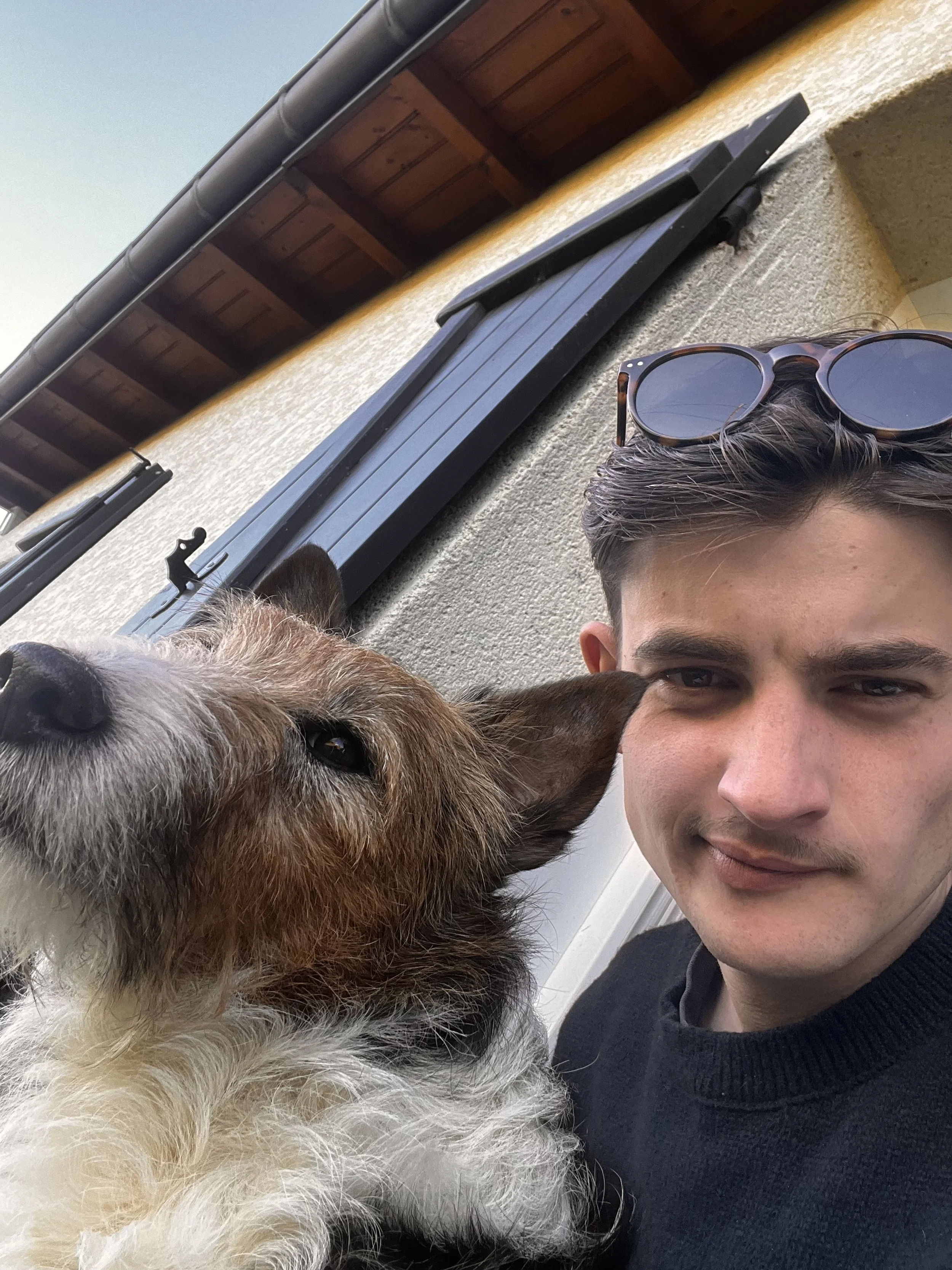 Un jeune homme avec des lunettes posées sur sa tête prend une selfie avec un chien de race indéterminée devant une maison avec volets noirs.