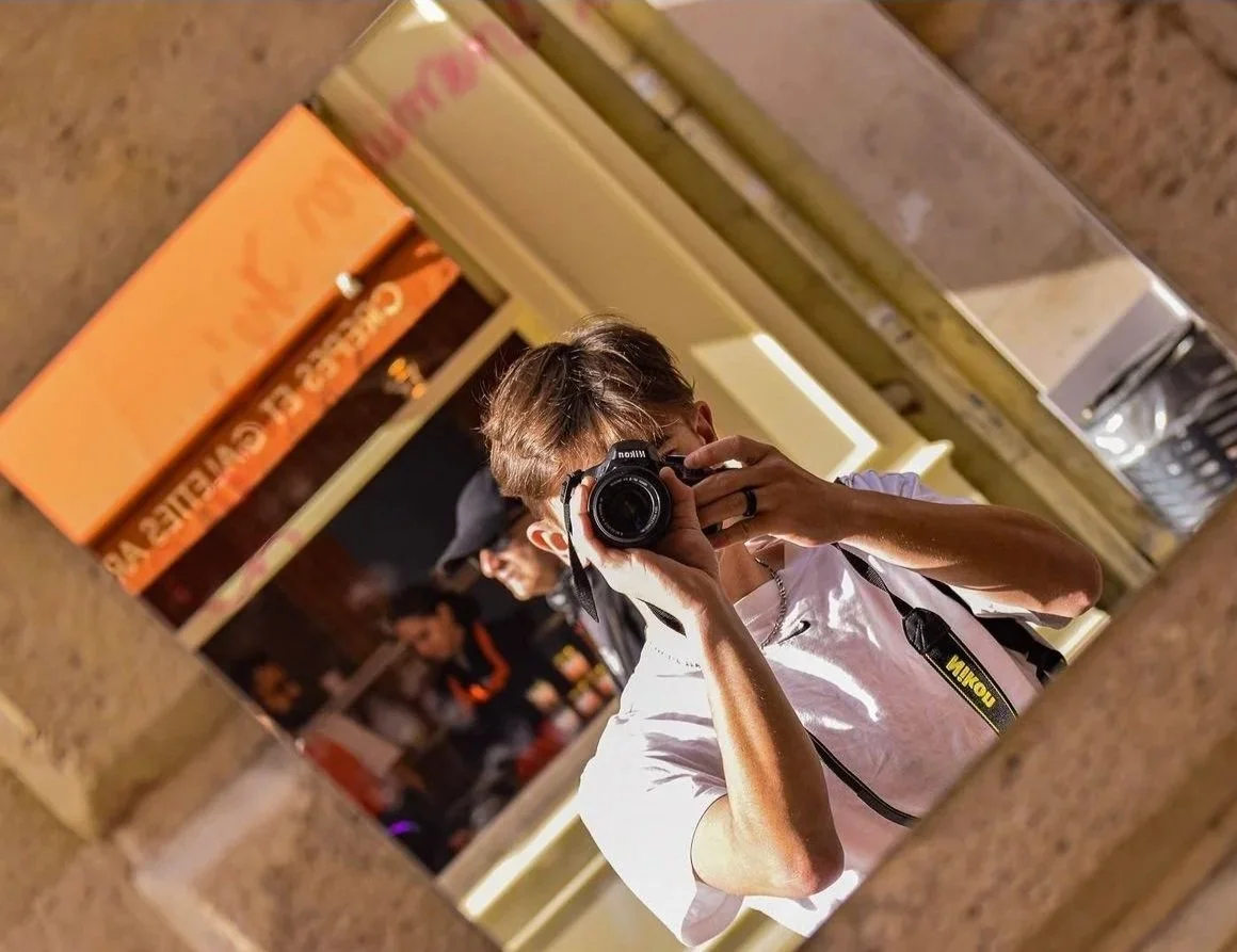 Un homme prend une photo de lui-même dans un miroir, portant un appareil photo Nikon avec un porte-caméra autour du cou, dans un environnement intérieur, avec un fond flou montrant des personnes et un panneau en anglais.