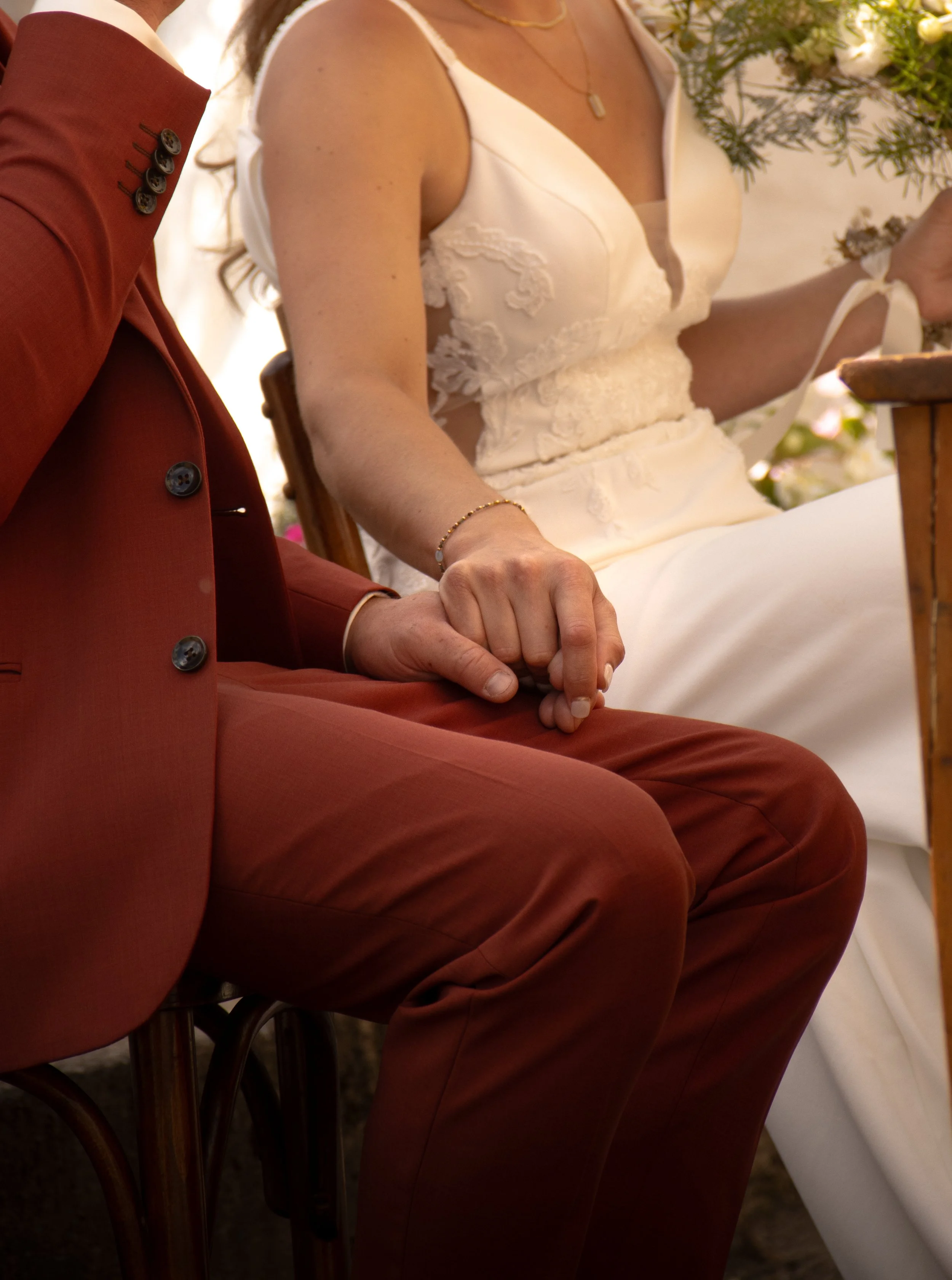 Deux personnes assises côte à côte, main dans la main, lors d'un mariage ou d'une cérémonie, l'une porte un costume rouge et l'autre une robe blanche.