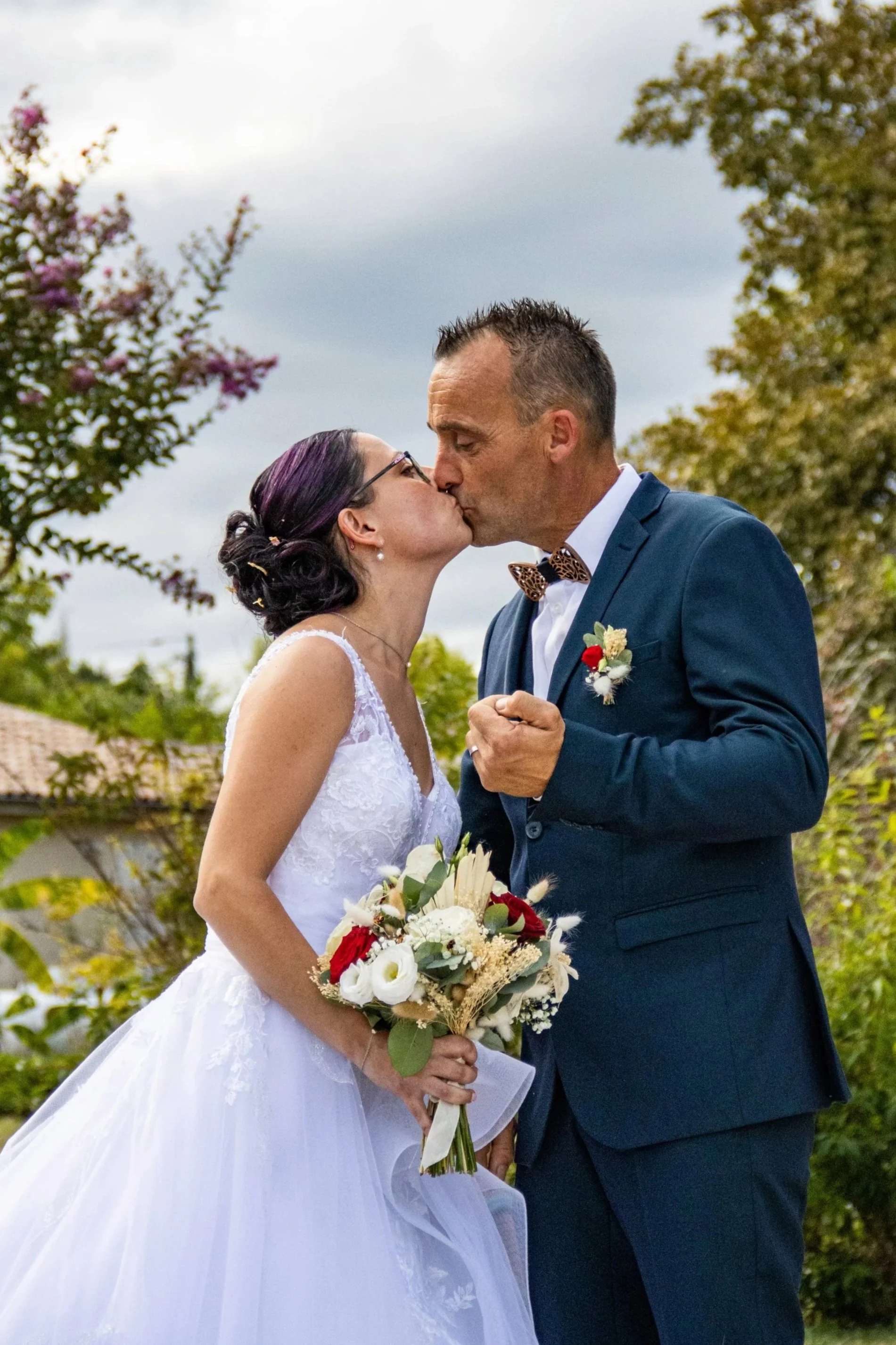 Un couple de mariés s'embrassent lors de leur mariage en extérieur, la femme portant une robe blanche et tenant un bouquet de fleurs, l'homme en costume bleu avec un nœud papillon à motif.