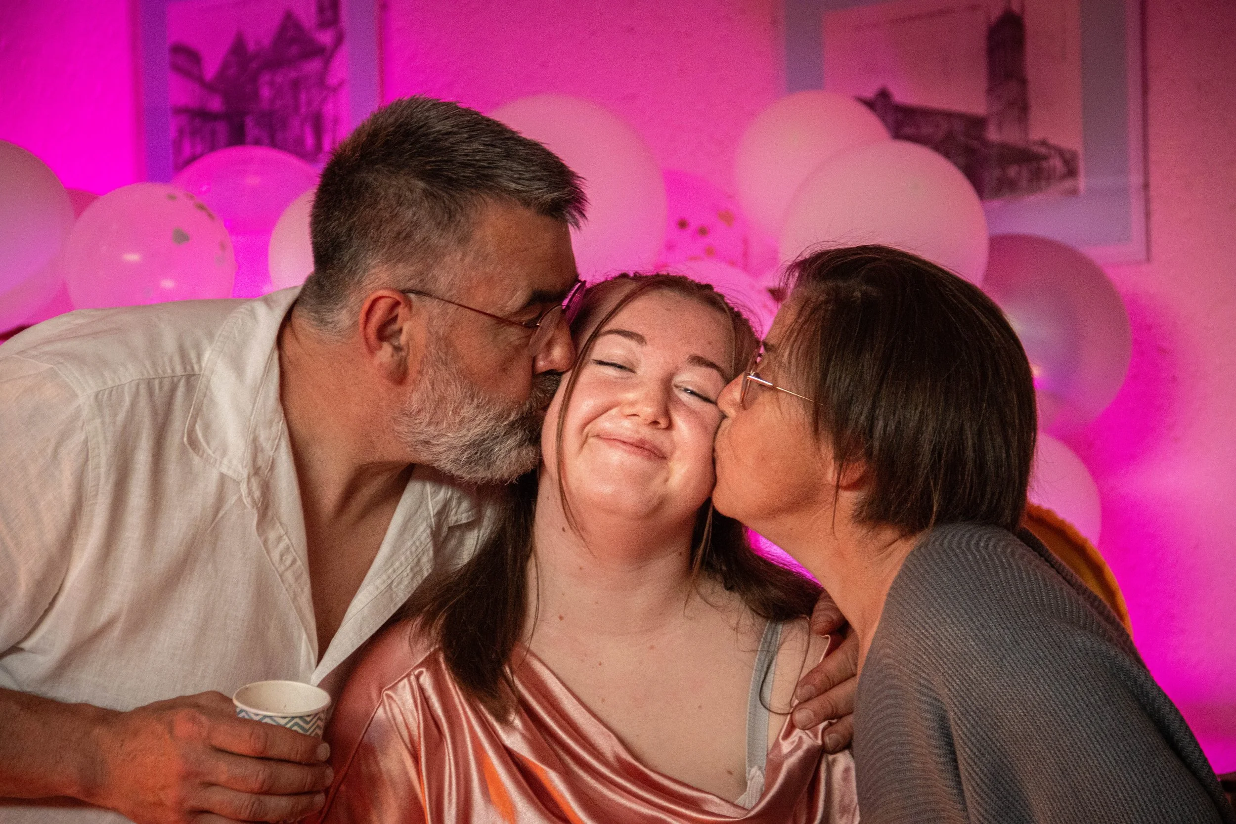 Une jeune femme souriante recevant deux bises, entourée de deux personnes âgées lors d'une fête avec décorations de ballons et lampions en arrière-plan.