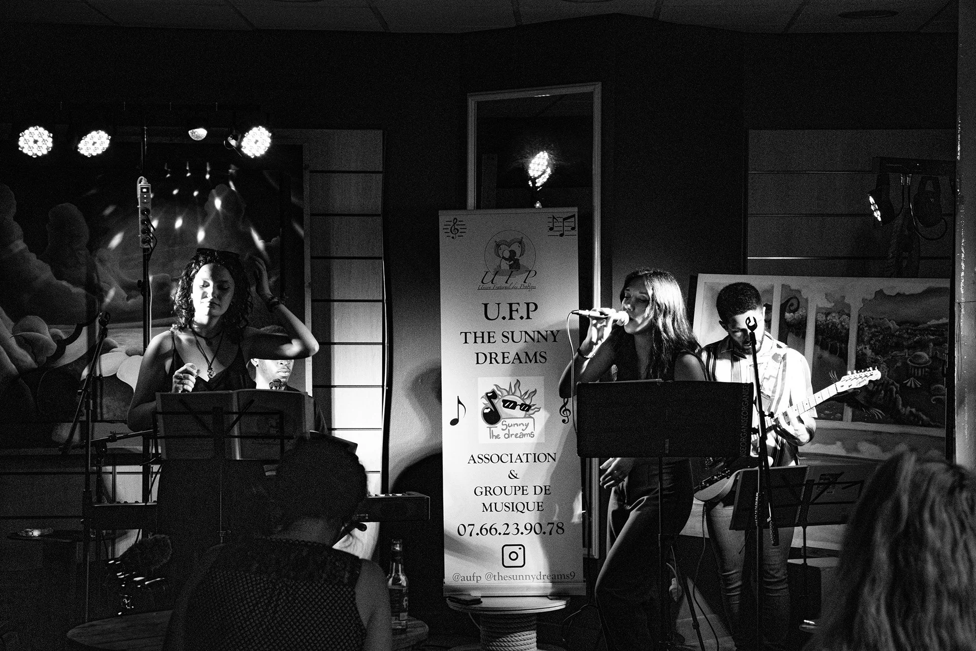 Groupe de musiciens en performance sur scène lors d'un concert, avec une chanteuse, un guitariste et un clavier. Décor sombre avec une affiche promotionnelle à l'arrière.