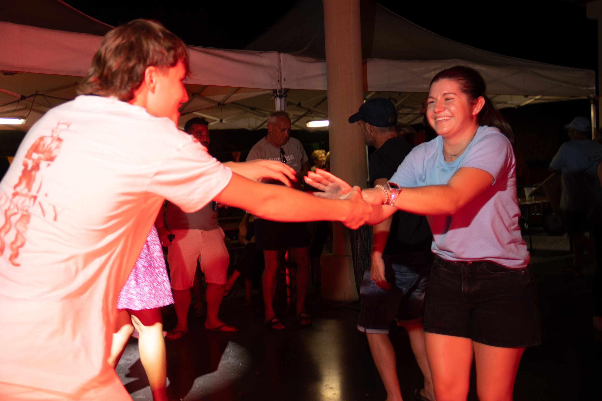 Deux personnes dansent lors d'une fête en plein air la nuit, souriant et tenant la main l'une de l'autre. Il y a d'autres personnes en arrière-plan sous une tente.
