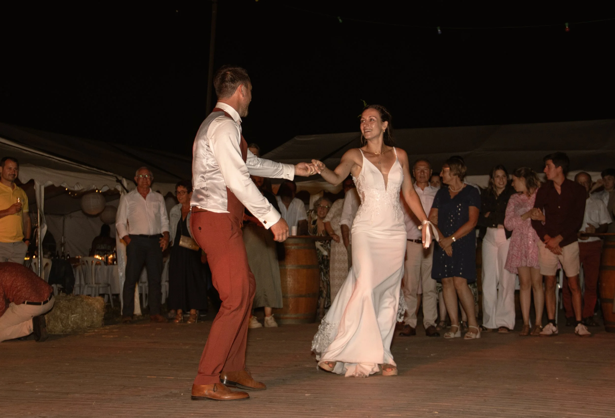 Un couple danse lors d'un mariage en soirée, entouré d'invités qui regardent et applaudissent. La mariée porte une robe blanche élégante, le marié est en pantalon marron et chemise blanche.