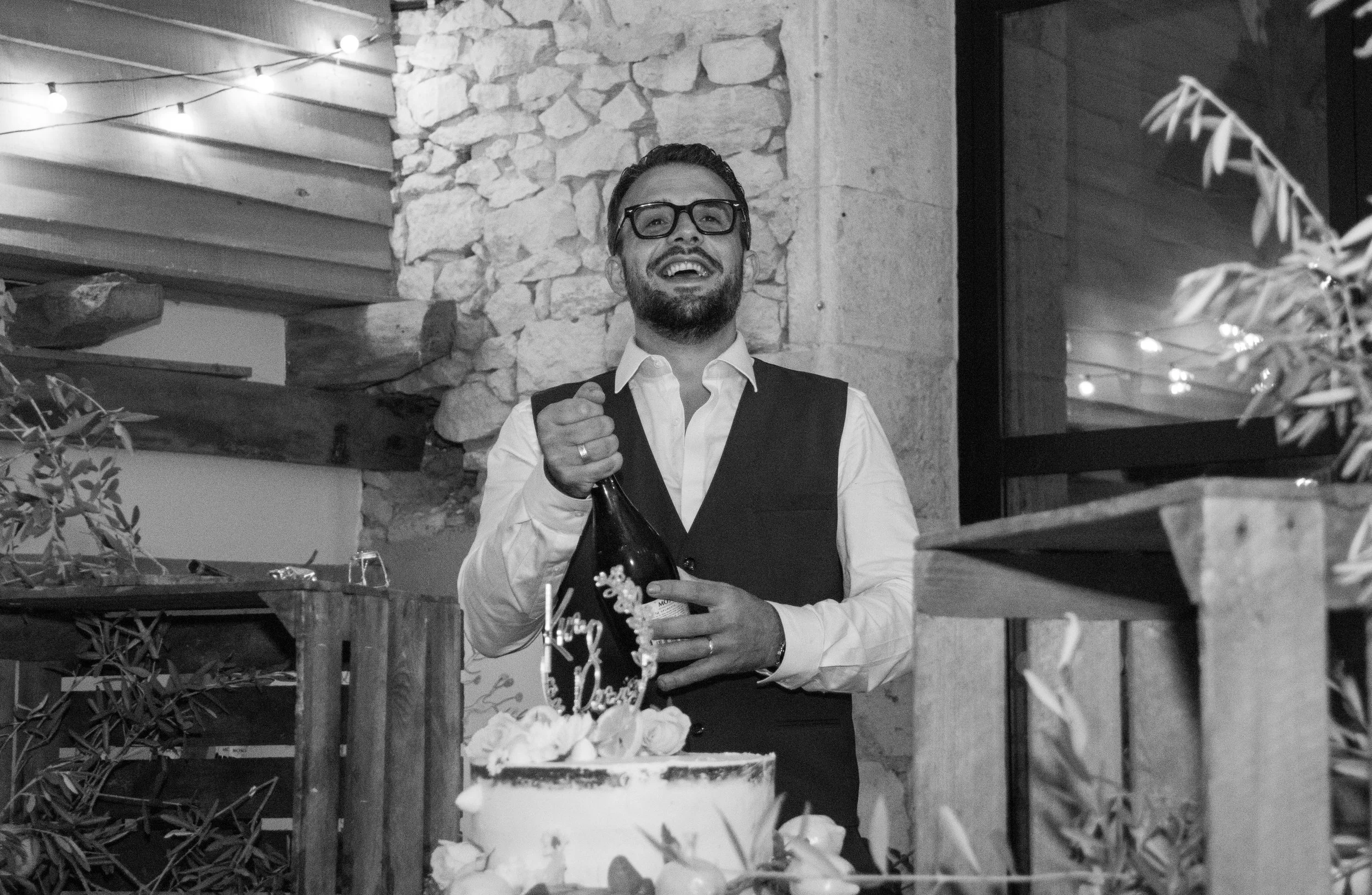 Un homme souriant avec des lunettes et une barbe, portant un gilet noir et une chemise blanche, tenant une bouteille de champagne lors d'une célébration d'anniversaire, avec un gâteau décoré devant lui dans un décor intérieur avec murs en pierre et décoration chaleureuse.