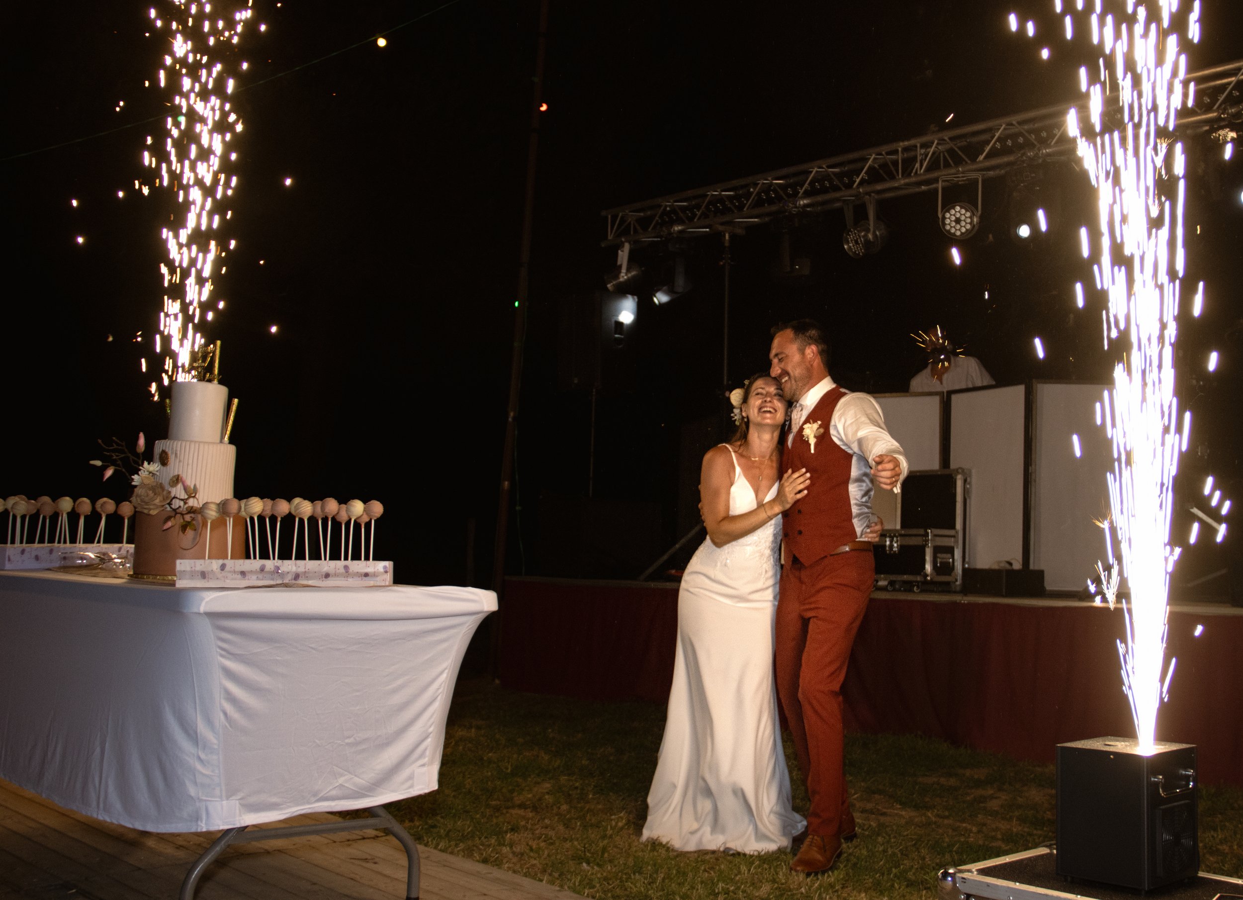 Un couple en robe de mariage danse lors d'une célébration, avec des feux d'artifice en arrière-plan et un gâteau de mariage à gauche.