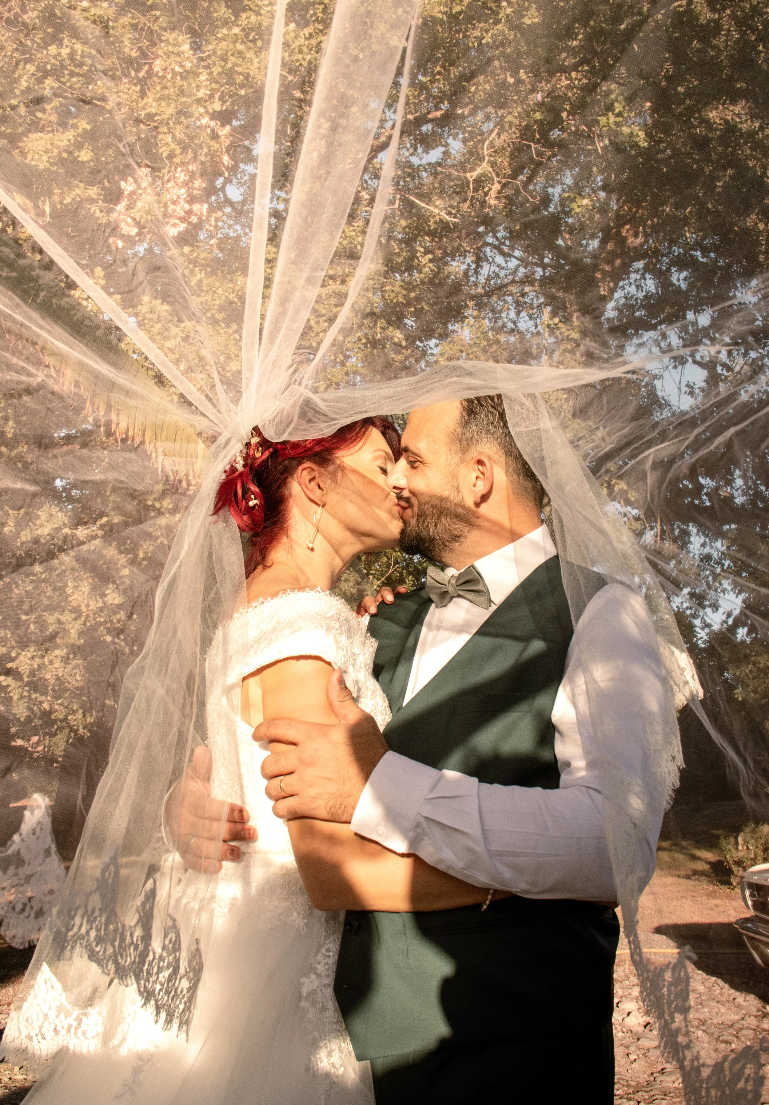 Un couple en mariage s'embrasse sous un voile, lors d'une séance photo en plein air avec des arbres en arrière-plan.