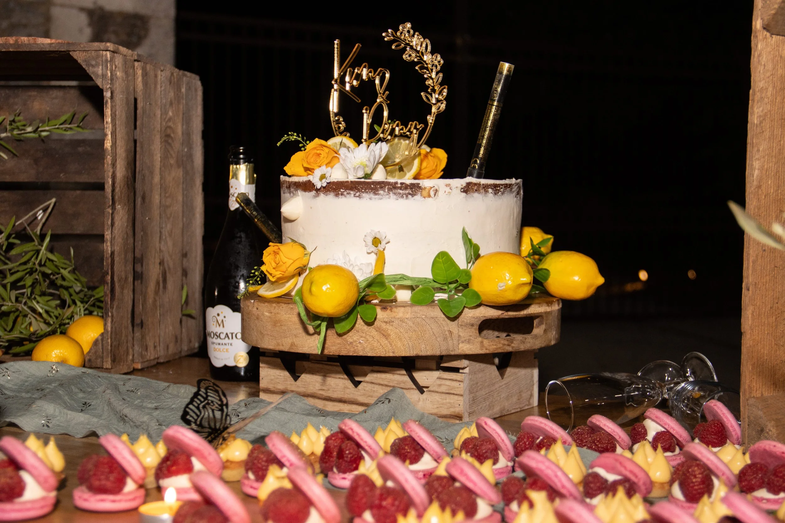Gâteau de mariage blanc décoré de fleurs jaunes et blanches, avec des citrons et des feuilles vertes sur un support en bois, entouré de macarons rose et jaunes avec des framboises, et une bouteille de soda à proximité.
