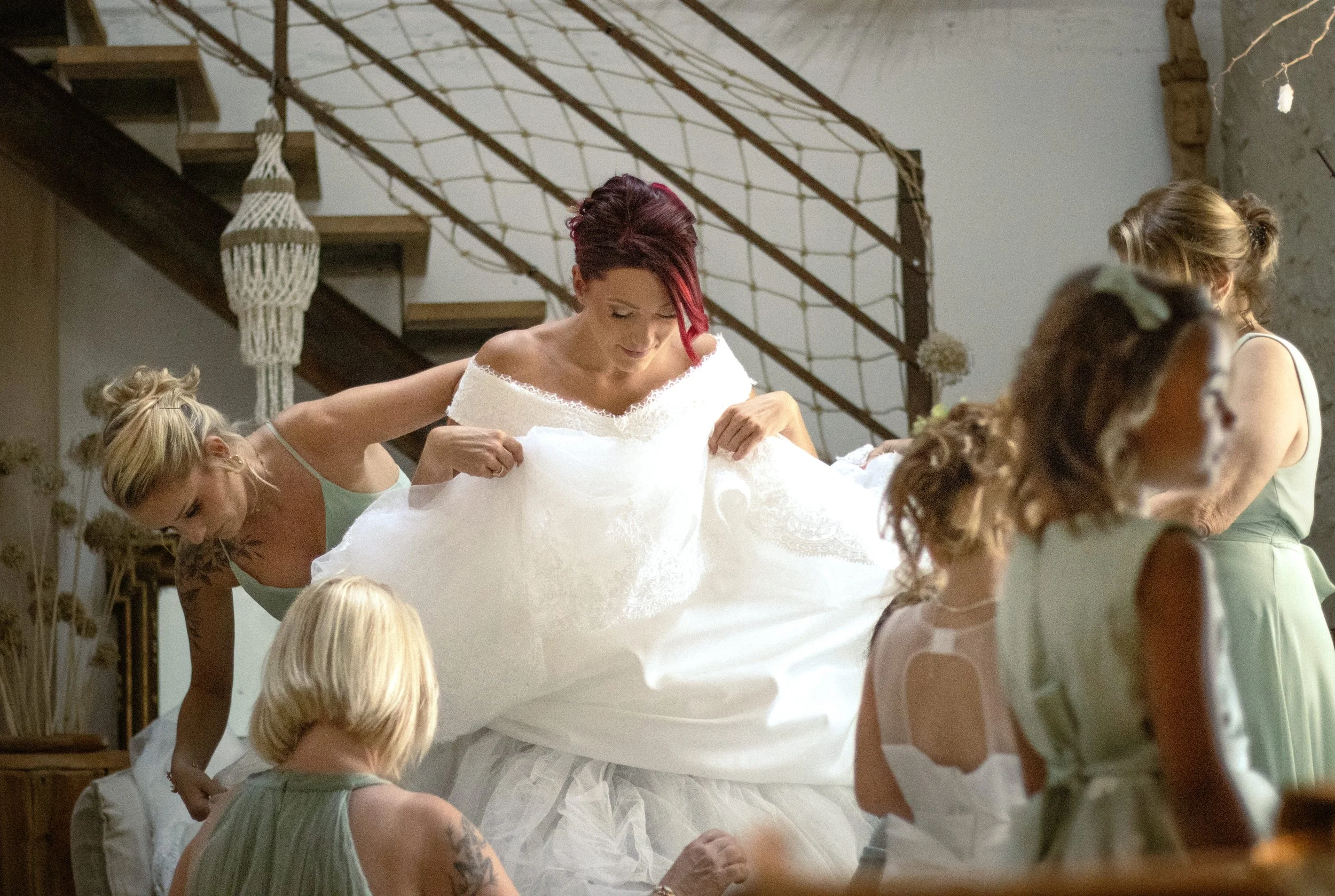 Une femme en robe de mariage entourée de plusieurs femmes qui l'aident à ajuster sa robe, dans une décoration intérieure chaleureuse.