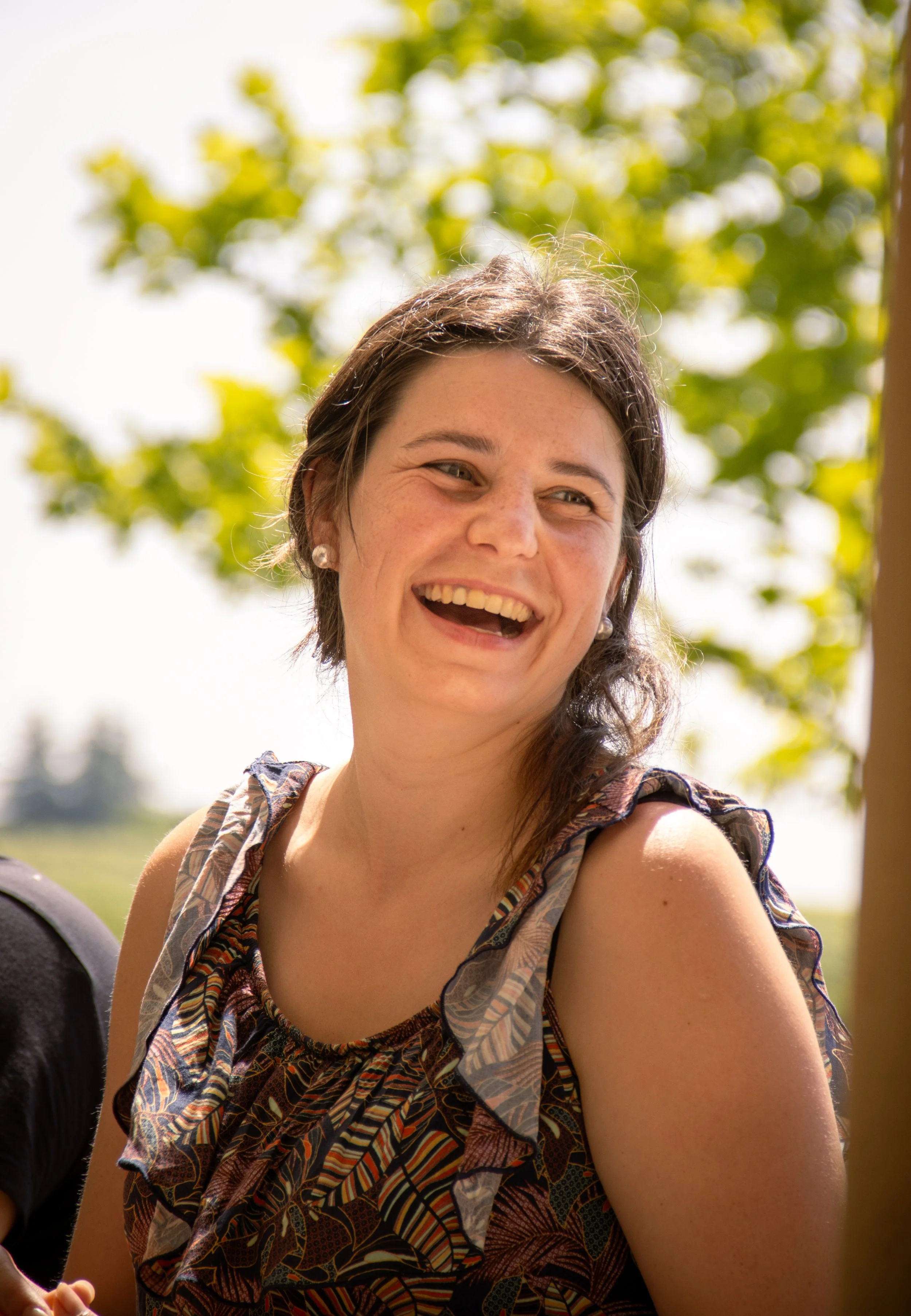 Une femme souriante avec des cheveux bruns, portant une robe avec un motif coloré, à l'extérieur avec des arbres verts et un ciel ensoleillé en arrière-plan.