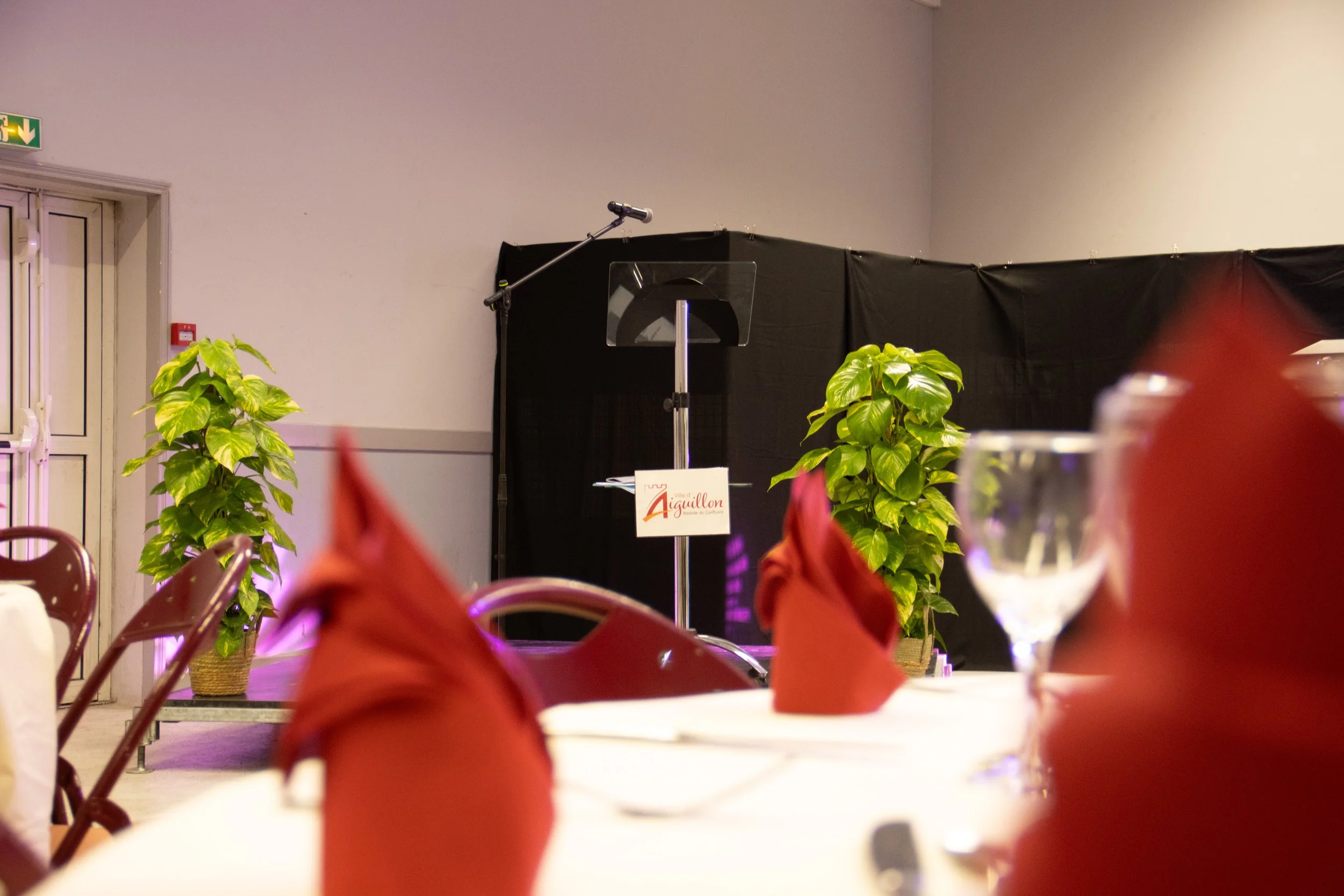 Une scène d'événement avec une scène vide, deux plantes vertes, des tables avec des nappes blanches et des serviettes pliées en forme de pyramide de couleur rouge, et un microphone sur un support. Il y a une porte avec une sortie de secours à gauche.