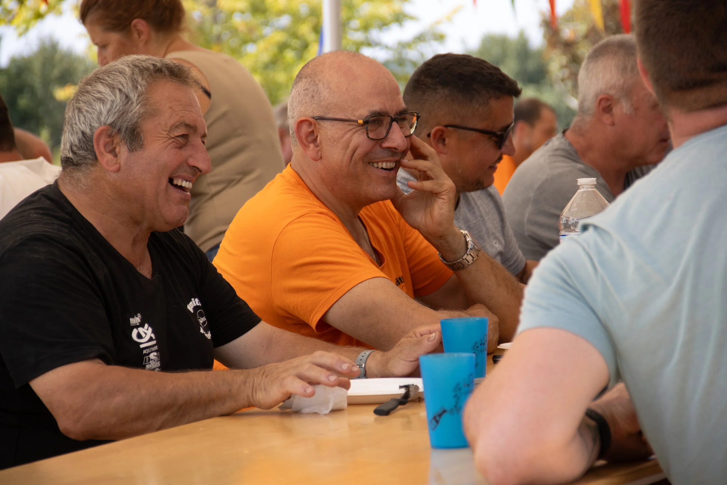 Groupe de personnes souriantes assises autour d'une table lors d'un événement en plein air.