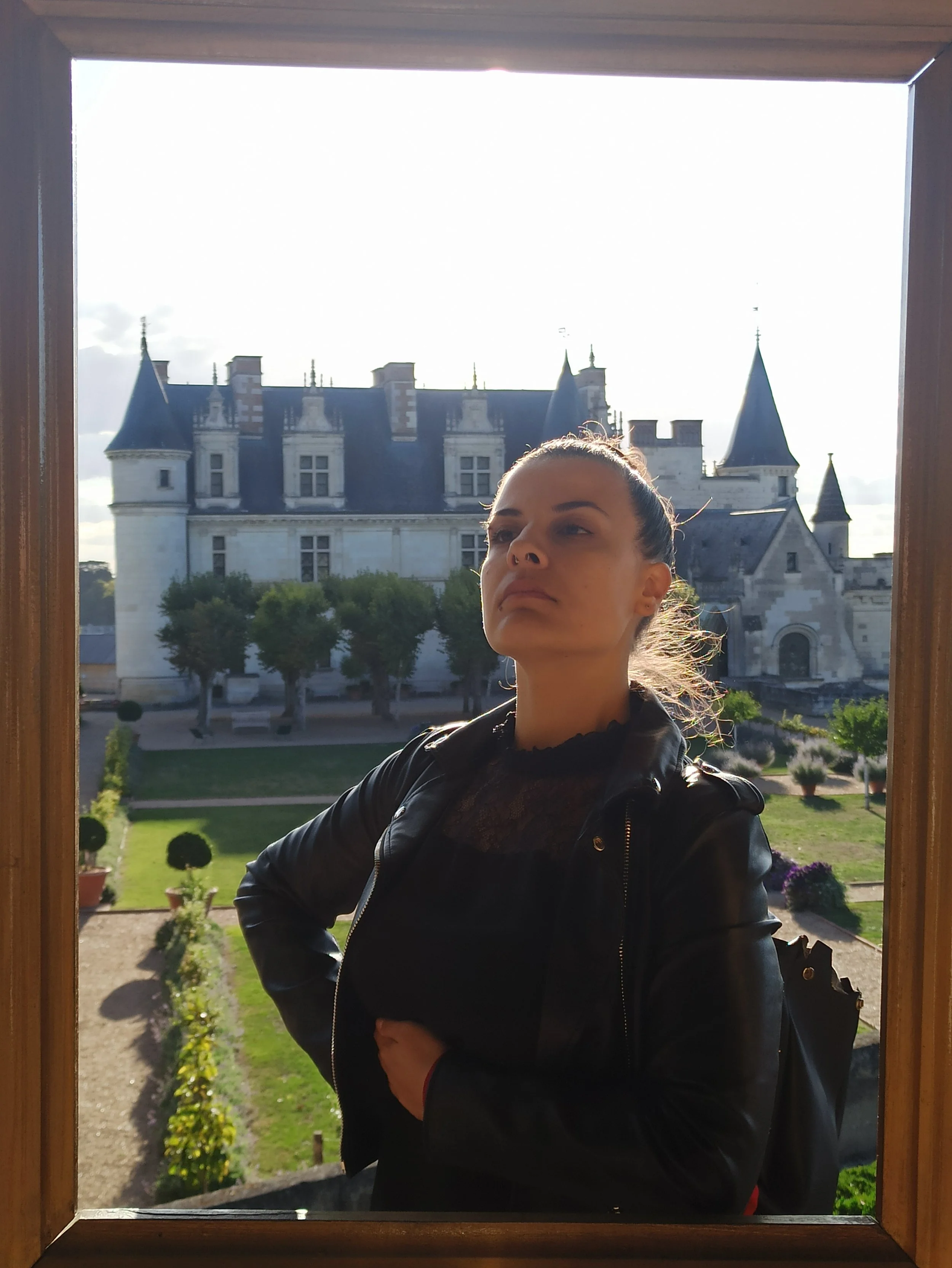 A woman wearing a black leather jacket standing outside near a castle, framed by a wooden border.