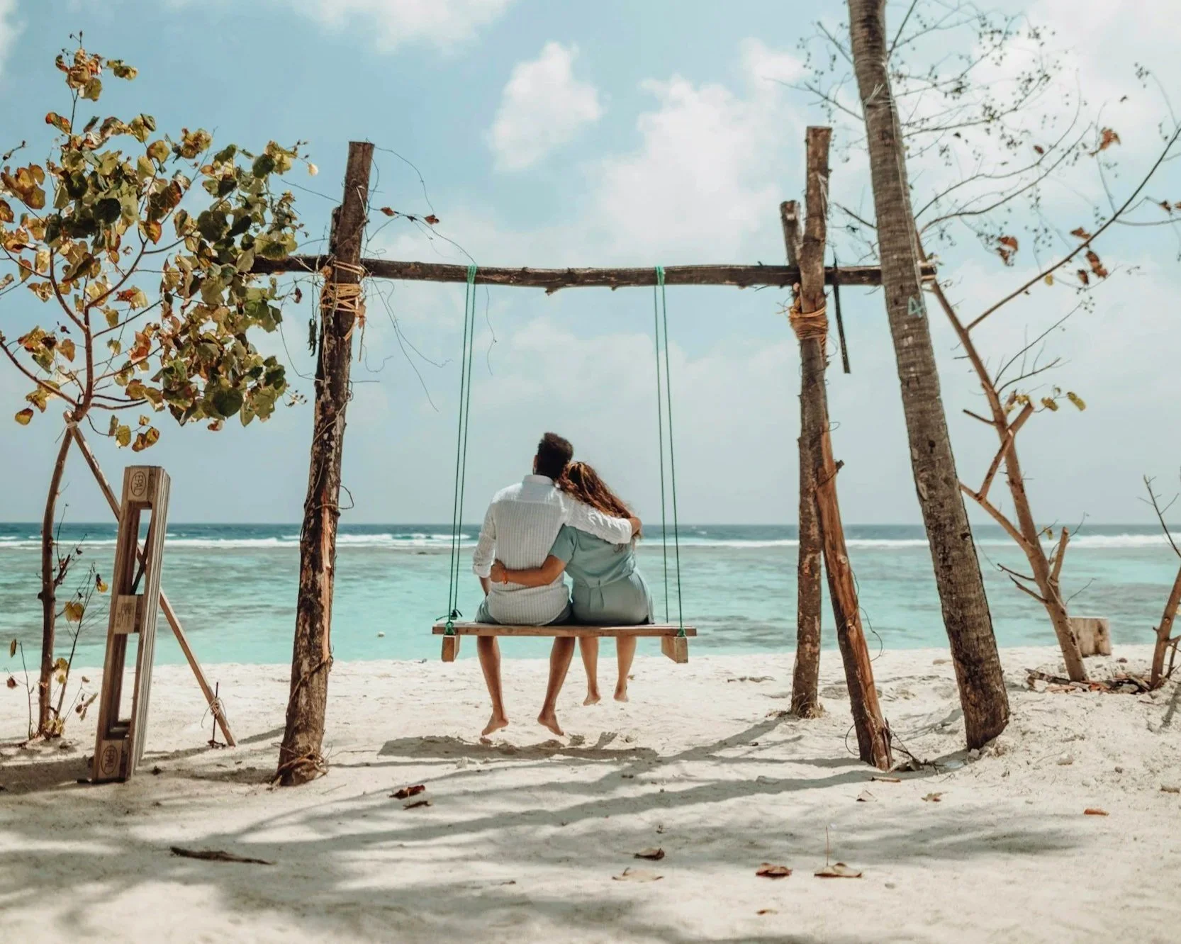 Un couple assis sur une balançoire en bois suspendue à une structure en bois sur une plage, regardant la mer.