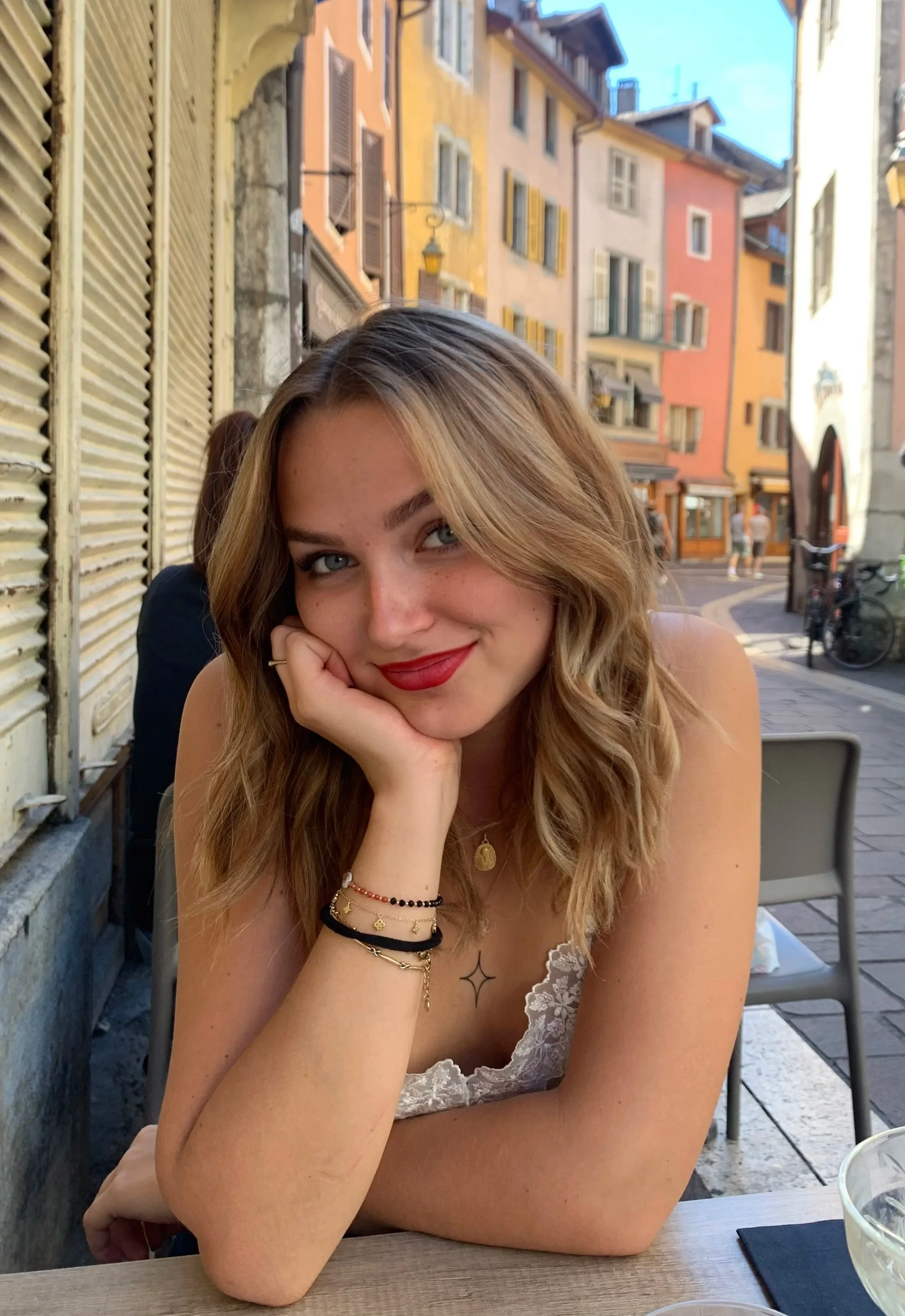 Une jeune femme aux cheveux bouclés, souriante, assise à une table en plein air dans une rue colorée, portant une robe blanche en dentelle et des bracelets.