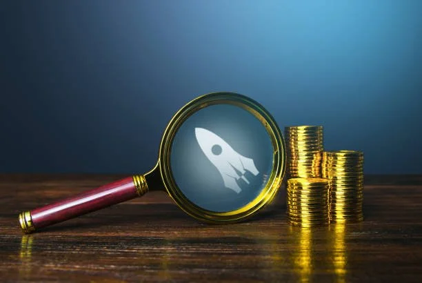 Magnifying glass with a rocket icon inside, placed next to stacks of gold coins on a wooden surface.