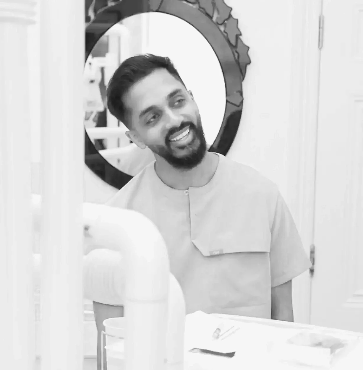 A man with a beard and short hair smiling and talking in front of a mirror in a salon or barbershop, with hair styling products and tools on the counter.