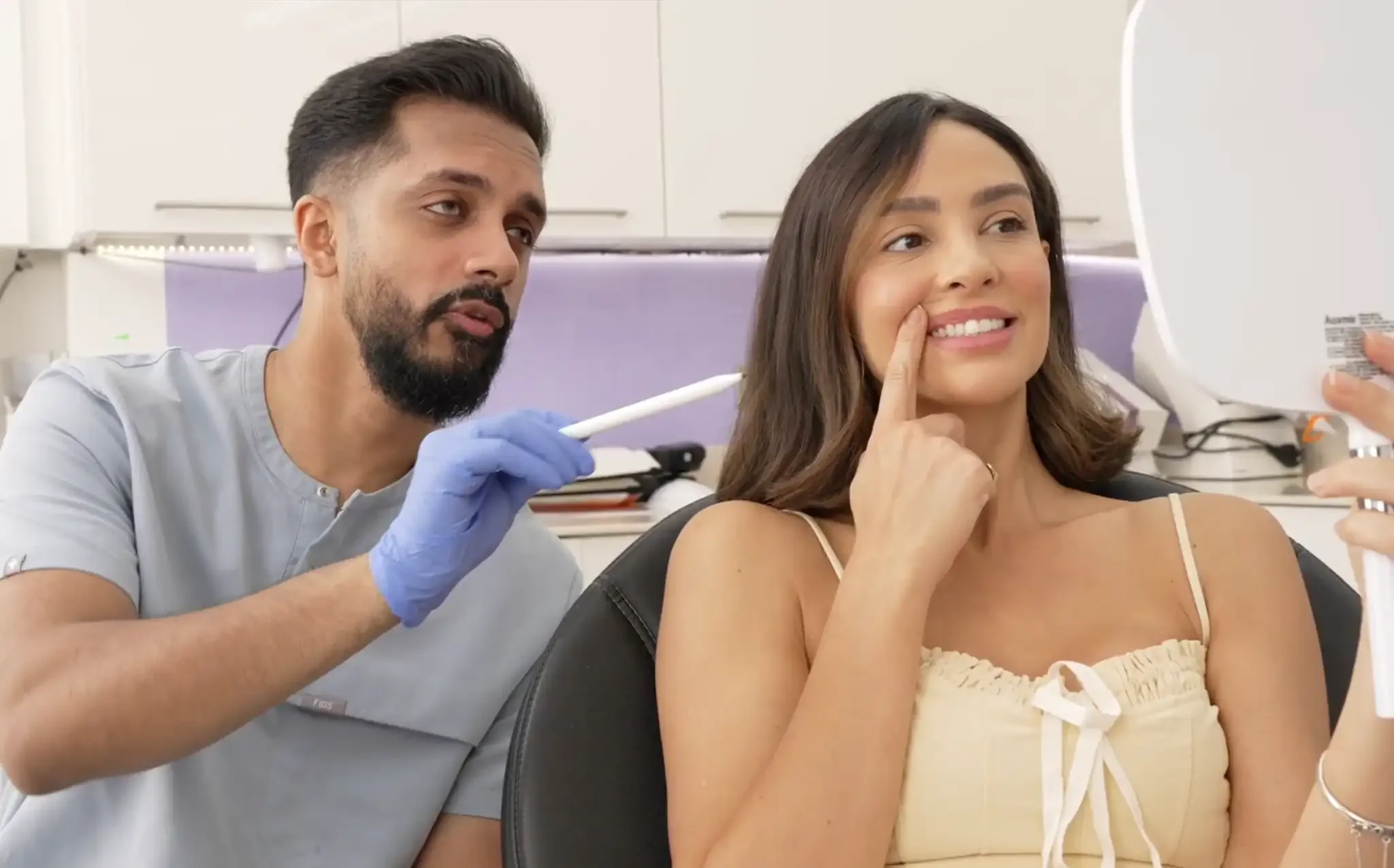 A woman at the dentist's office smiling and pointing at her cheek while looking at her phone, with a dentist preparing to take a swab sample.