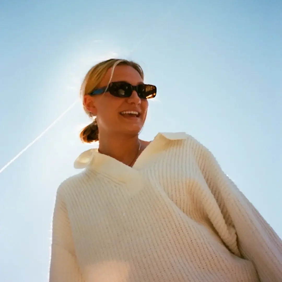 A woman wearing sunglasses and a white knit sweater smiling against a clear blue sky.