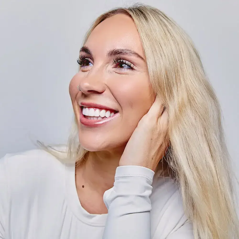 Close-up of a smiling woman with long blonde hair, wearing a white top, touching her hair, against a plain background.