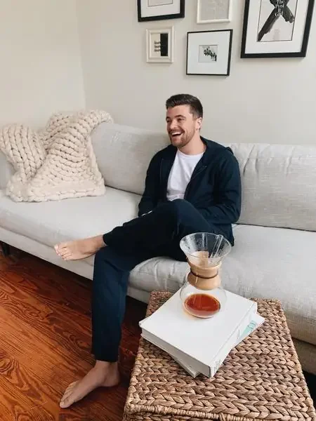 A man sitting on a white sofa, smiling, with a beige knit blanket, in a living room with framed wall art, a coffee table with a Chemex coffee maker, a glass of coffee, and books.