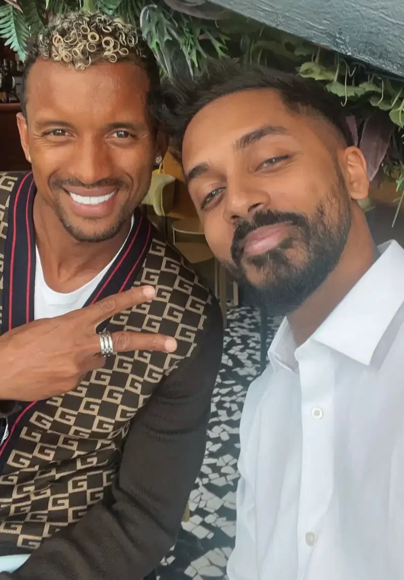 Two men smiling and taking a selfie at a restaurant or cafe, with green plants in the background.