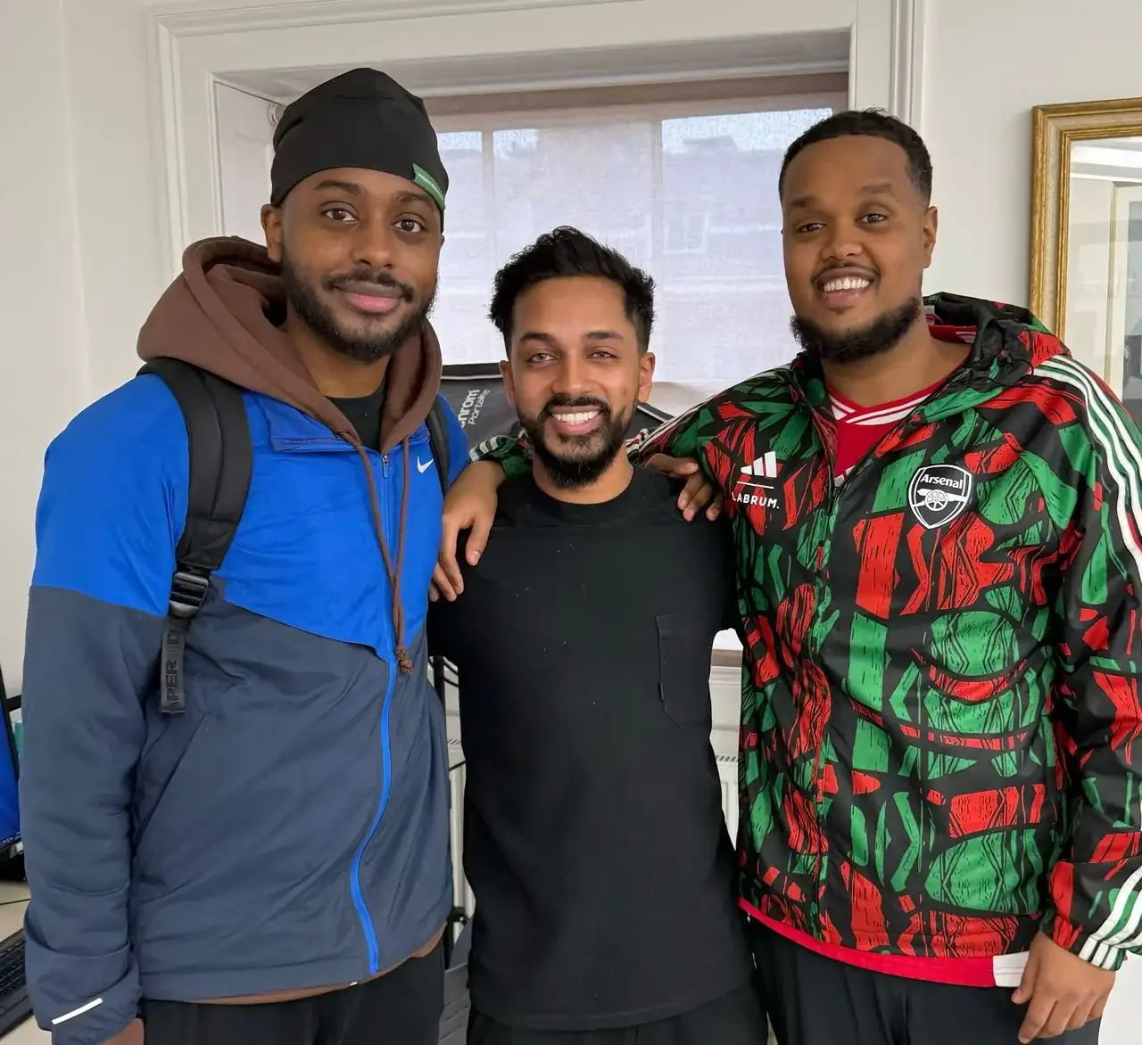 Three men smiling and standing close together indoors, with arms around each other's shoulders.