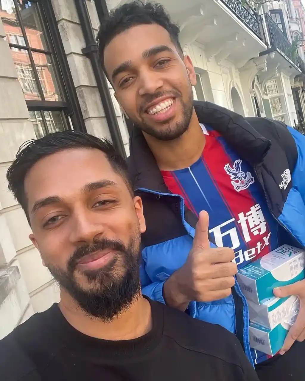 Two men taking a selfie in an urban neighborhood. One man has a beard and is wearing a black shirt, and the other man is smiling, giving a thumbs up, and wearing a colorful sports jersey with a jacket.