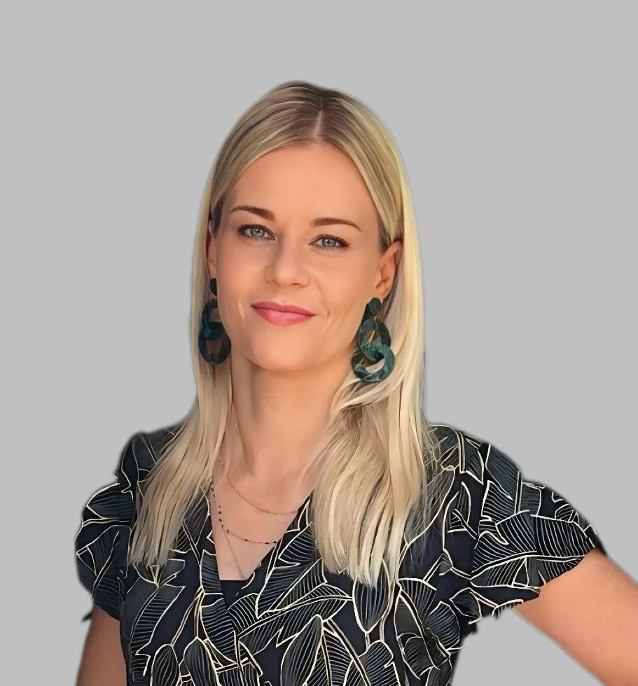Eine Frau mit langen blonden Haaren trägt große, dunkel gestreifte Ohrringe und ein schwarzes Kleid mit weißen Blattmustern. Sie lächelt leicht und steht vor einem neutral grauen Hintergrund.
