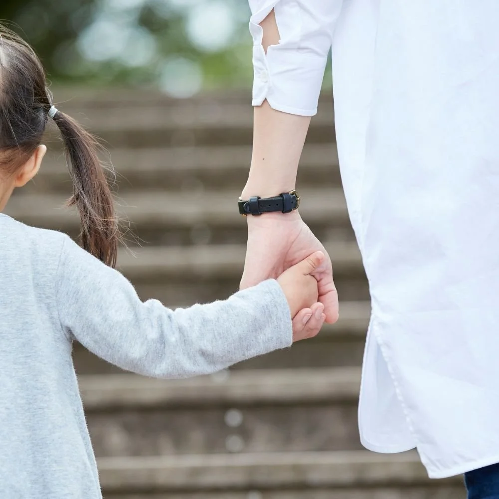 Ein Erwachsener hält die Hand eines Kindes. Der Hintergrund zeigt eine Treppe aus Holz. Der Erwachsene trägt ein weißes Hemd mit aufgerollten Ärmeln und einer schwarzen Uhr.