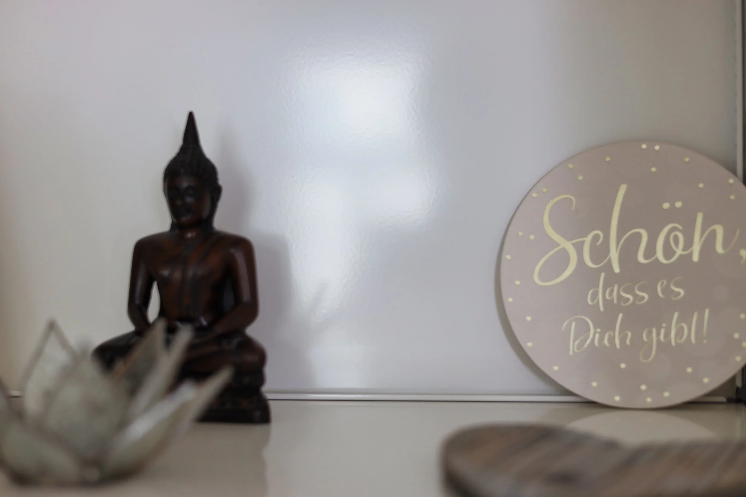 Deko-Szene mit einer kleinen Buddha-Statue, einem Keramik-Relief und einem runden Spiegel mit Aufschrift in deutscher Sprache.