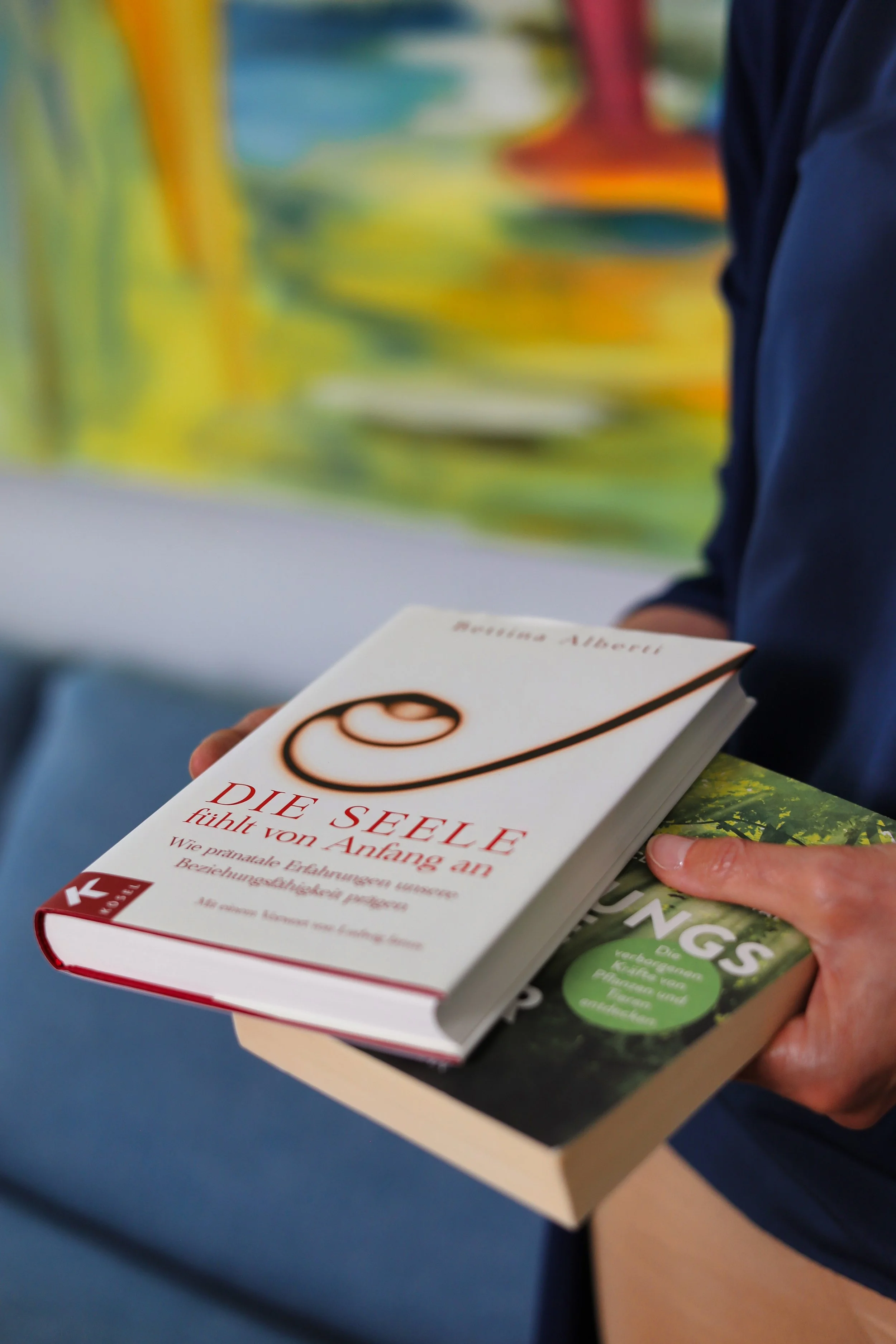Eine Person hält zwei Bücher in den Händen, ein offenes Buch mit dem Titel 'DIE SEELE fühlt von Anfang an' und ein weiteres Buch darunter. Der Hintergrund ist unscharf mit bunten, abstrakten Mustern.